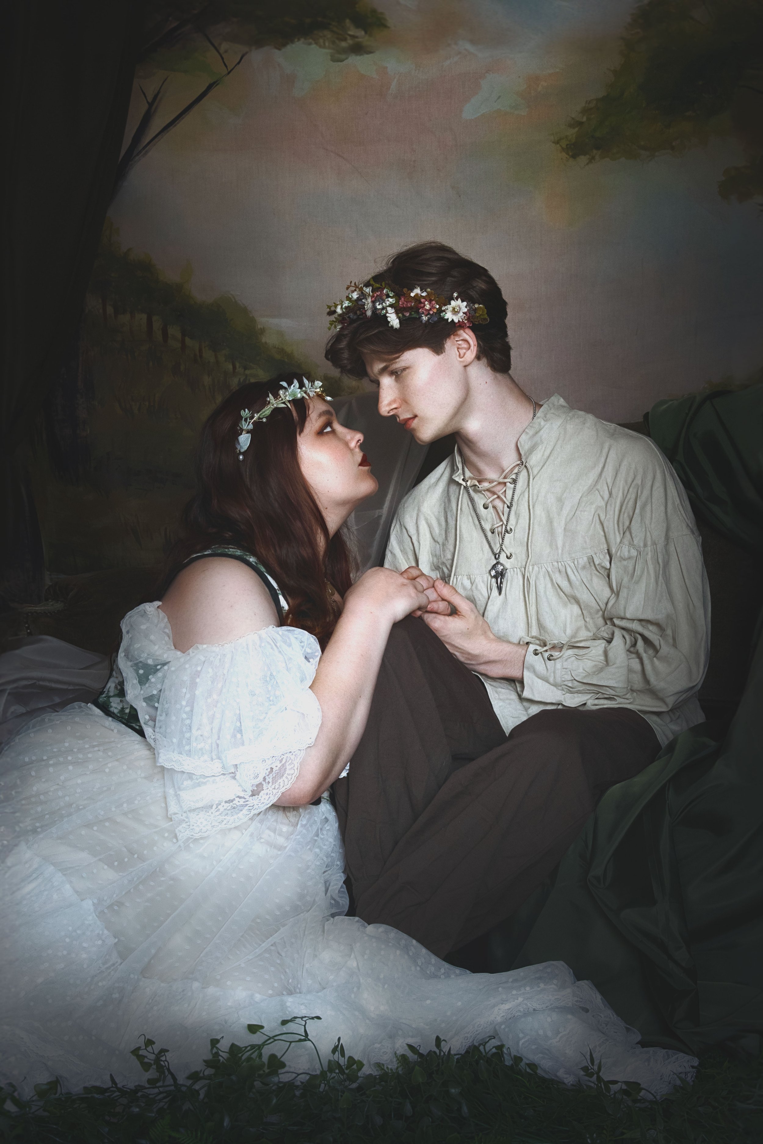 A romantic scene with a woman in a white lace dress and a man in medieval-style clothing, wearing floral crowns, sitting close and holding hands.