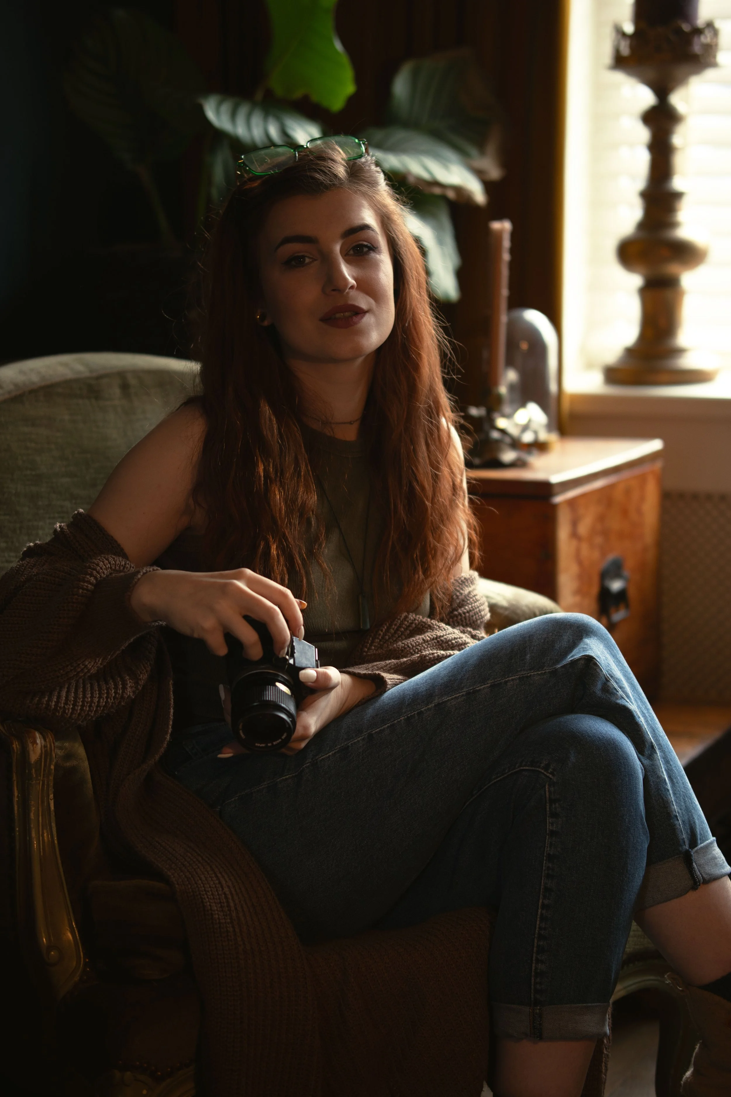 Woman with long hair sitting on an armchair holding a camera, indoor setting.