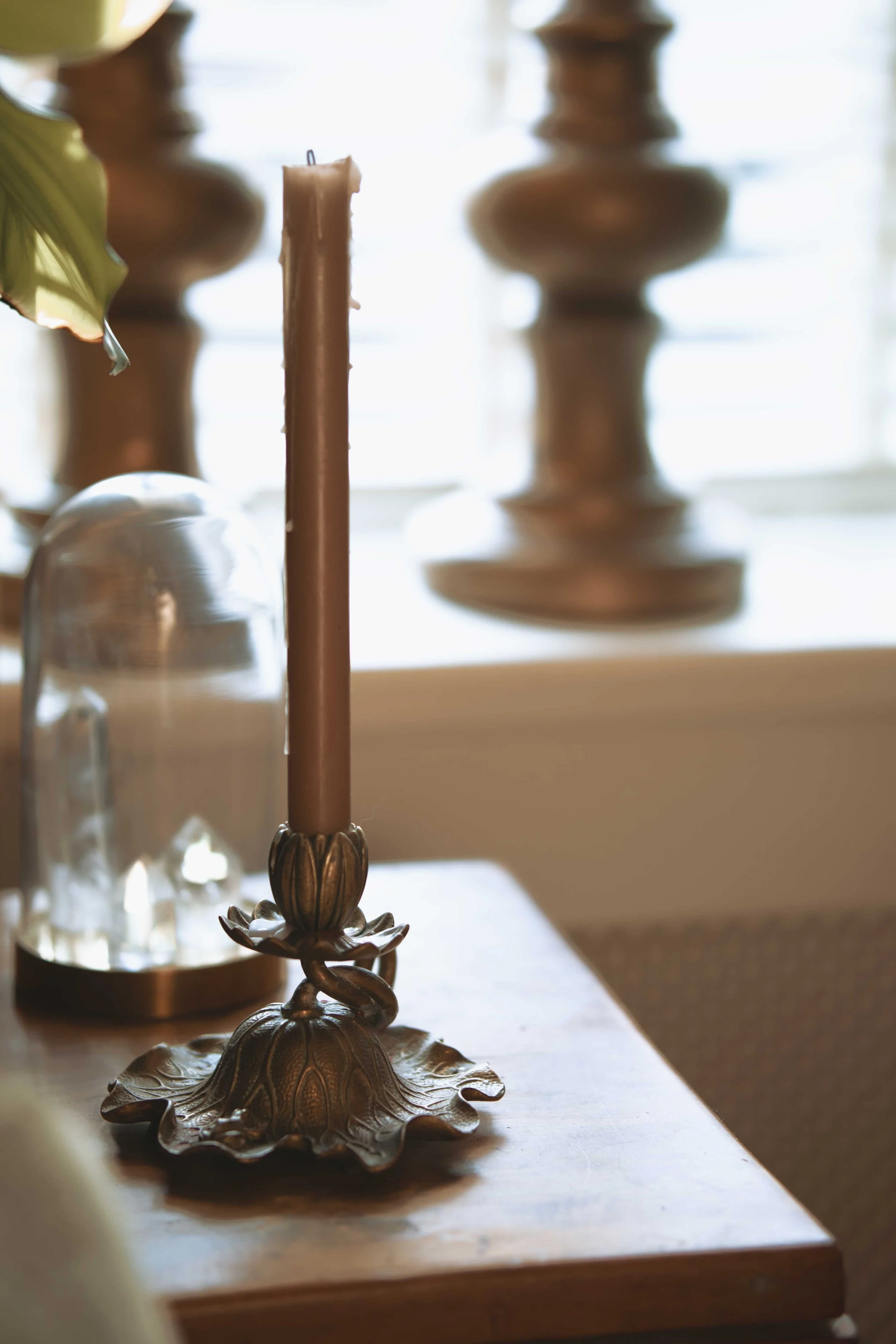 Decorative candleholder with an ornate design holding a brown candle on a wooden table; blurred background with a glass dome and abstract shapes.