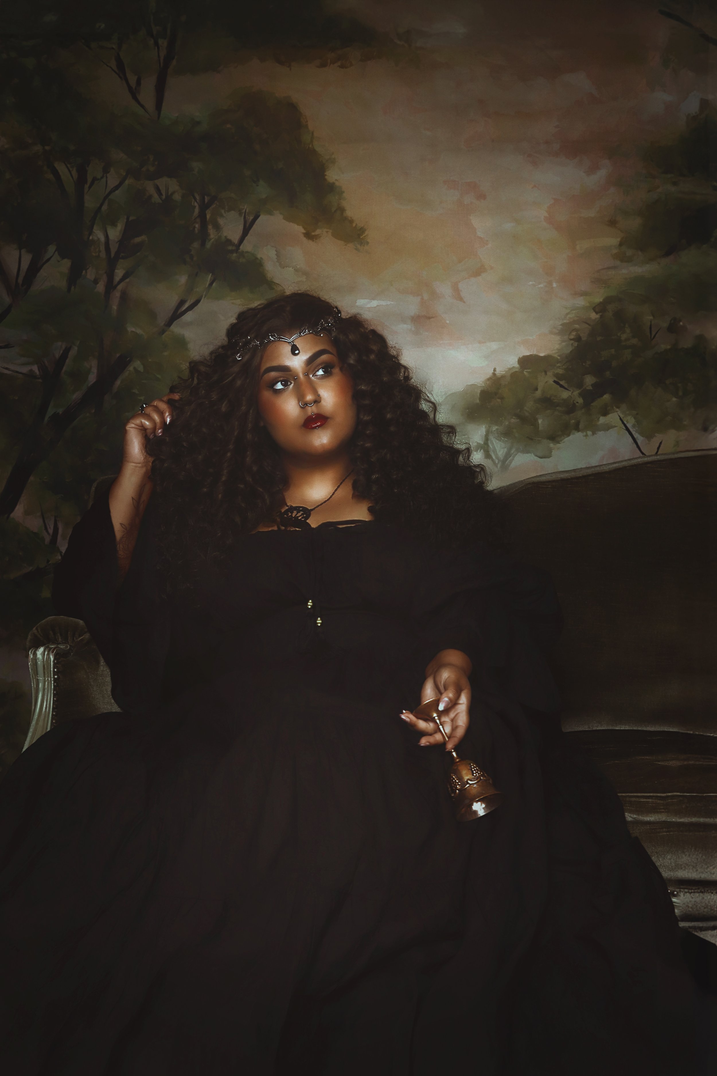 Person in dark attire with curly hair sitting on a vintage sofa, holding a decorative cup, against a background of painted trees.