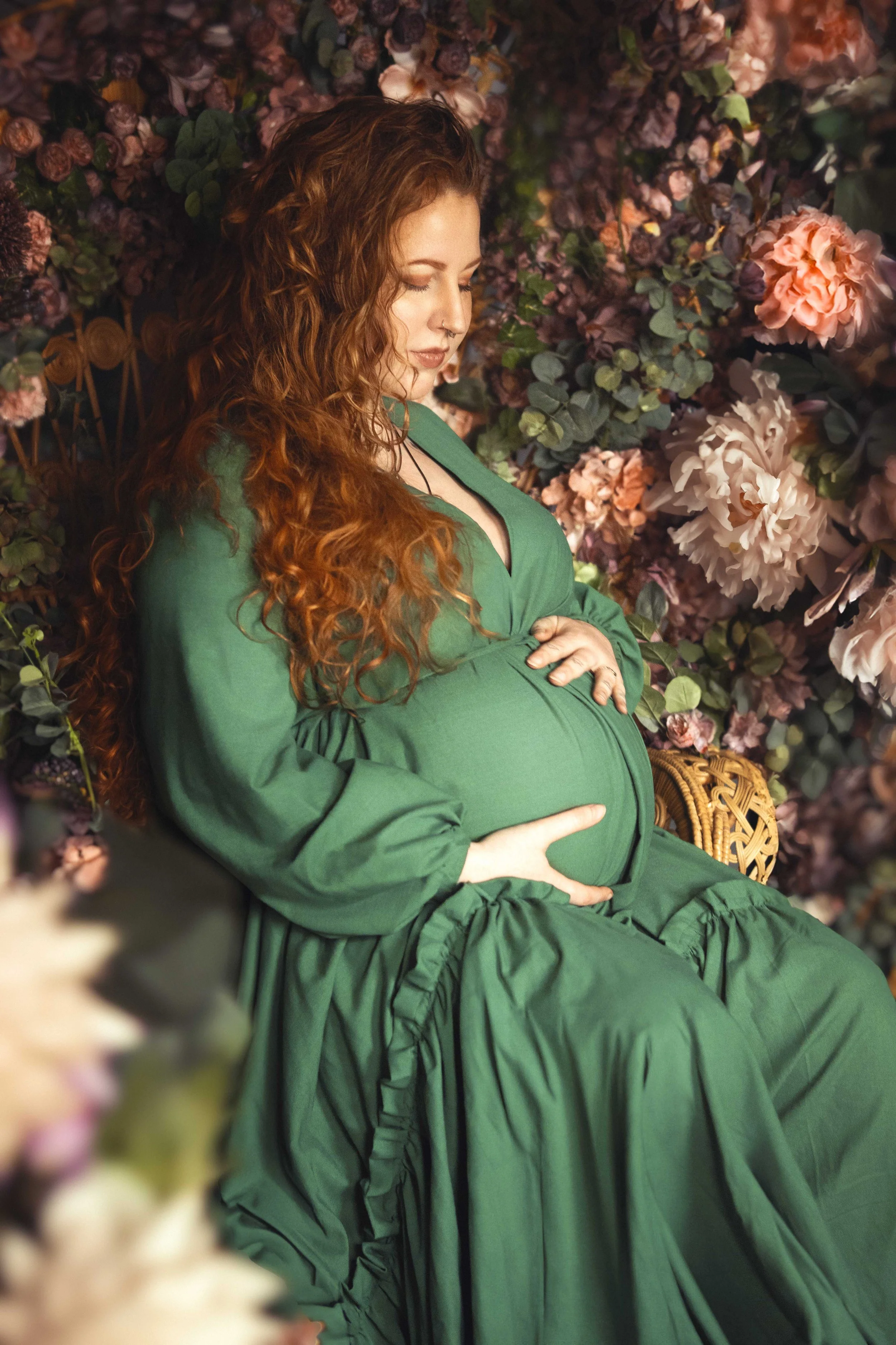 Pregnant woman in green dress seated against floral background, holding her belly.