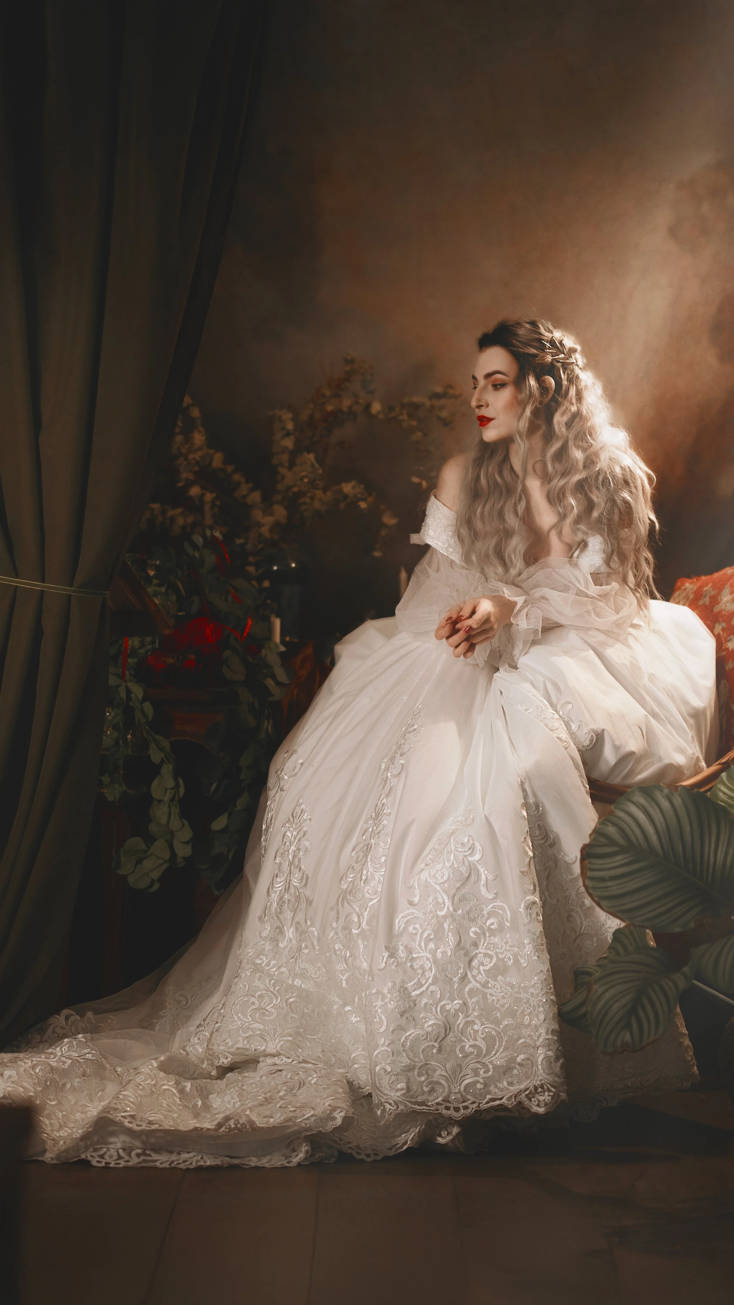 A woman in an elaborate white lace dress sitting thoughtfully in a dimly lit room with plants in the foreground and background.