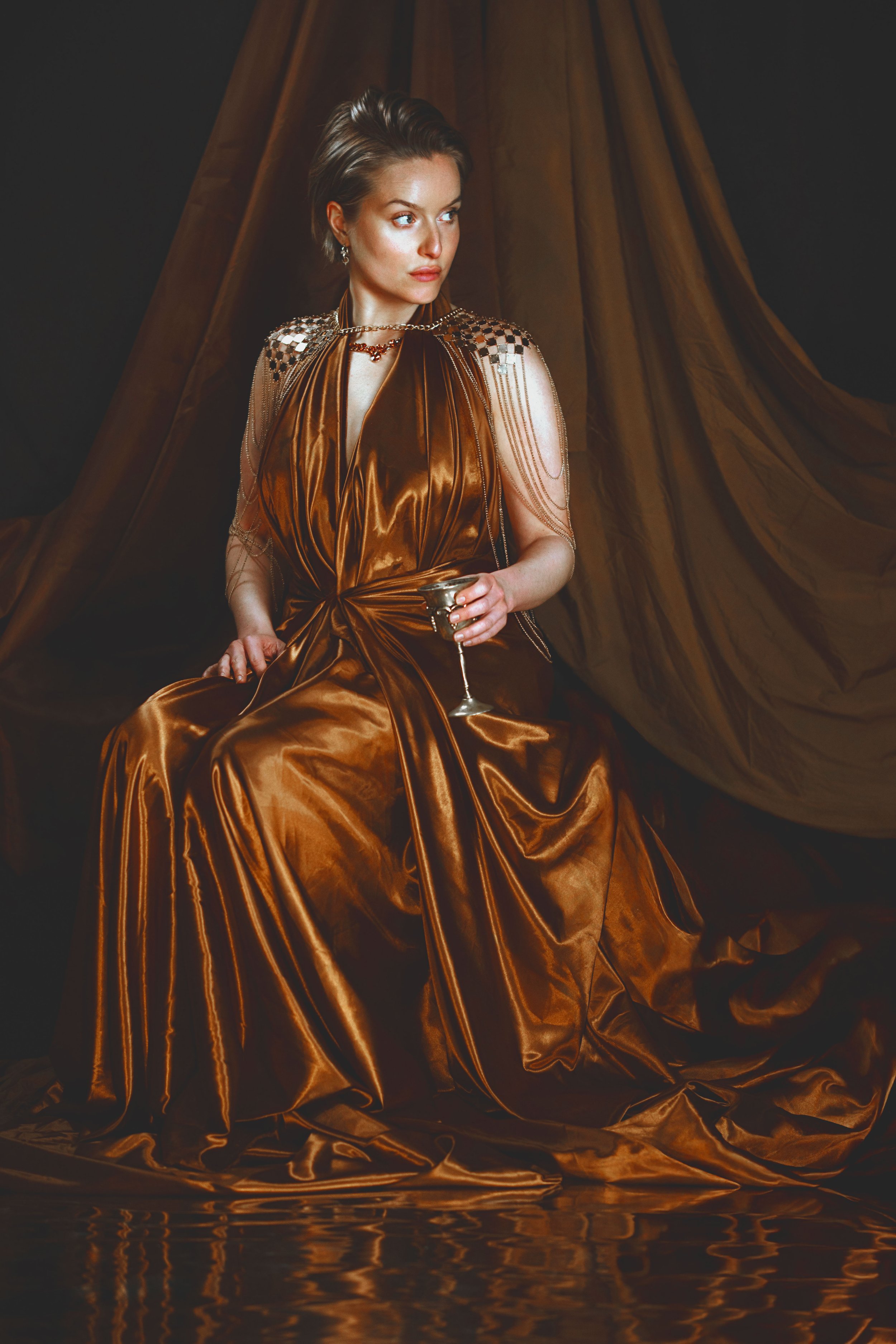 Woman in a luxurious satin gown holding a wine glass, with a dramatic draped brown fabric background.