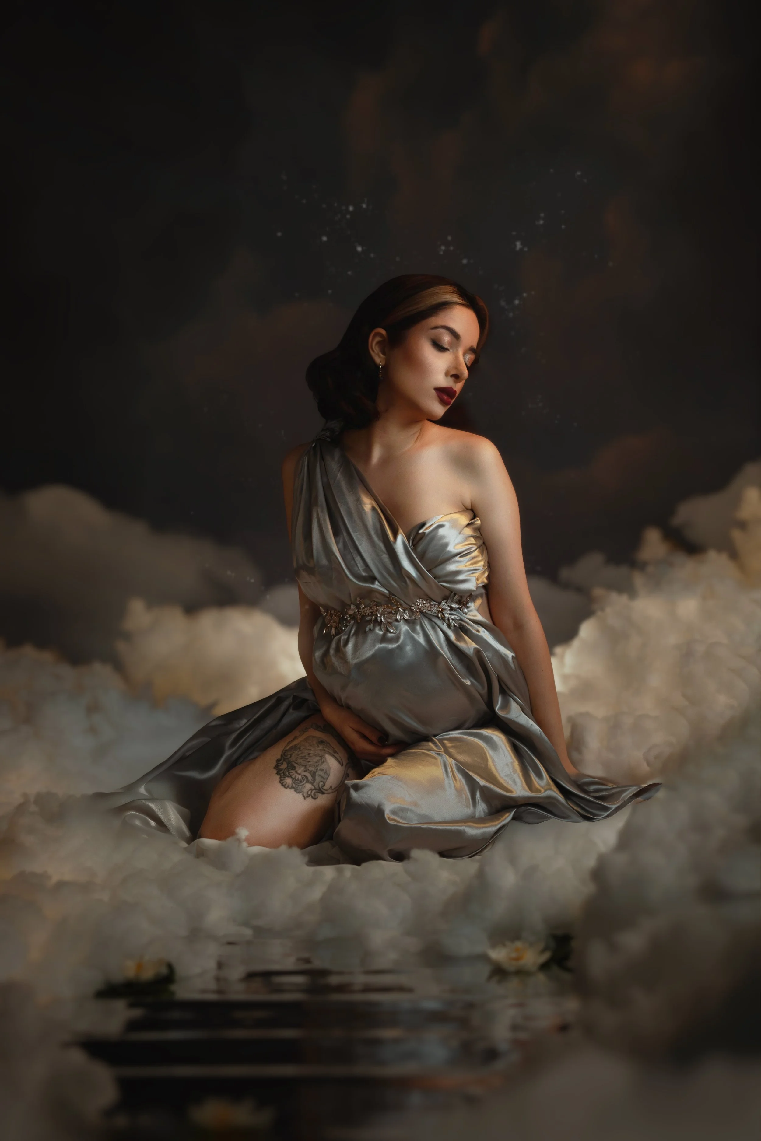Pregnant woman in a flowing silver dress sitting on clouds, holding her belly, with a dark, starry background.