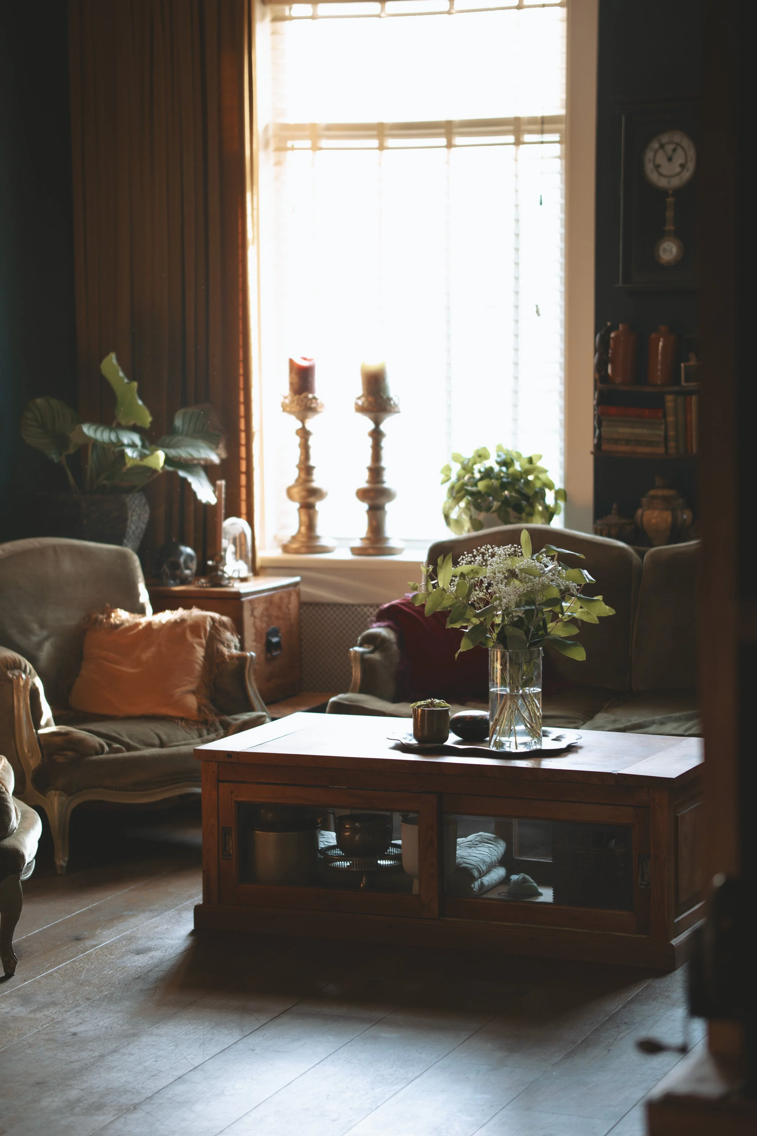Cozy living room with vintage armchairs, wooden coffee table, potted plants, and decorative candles near a window.