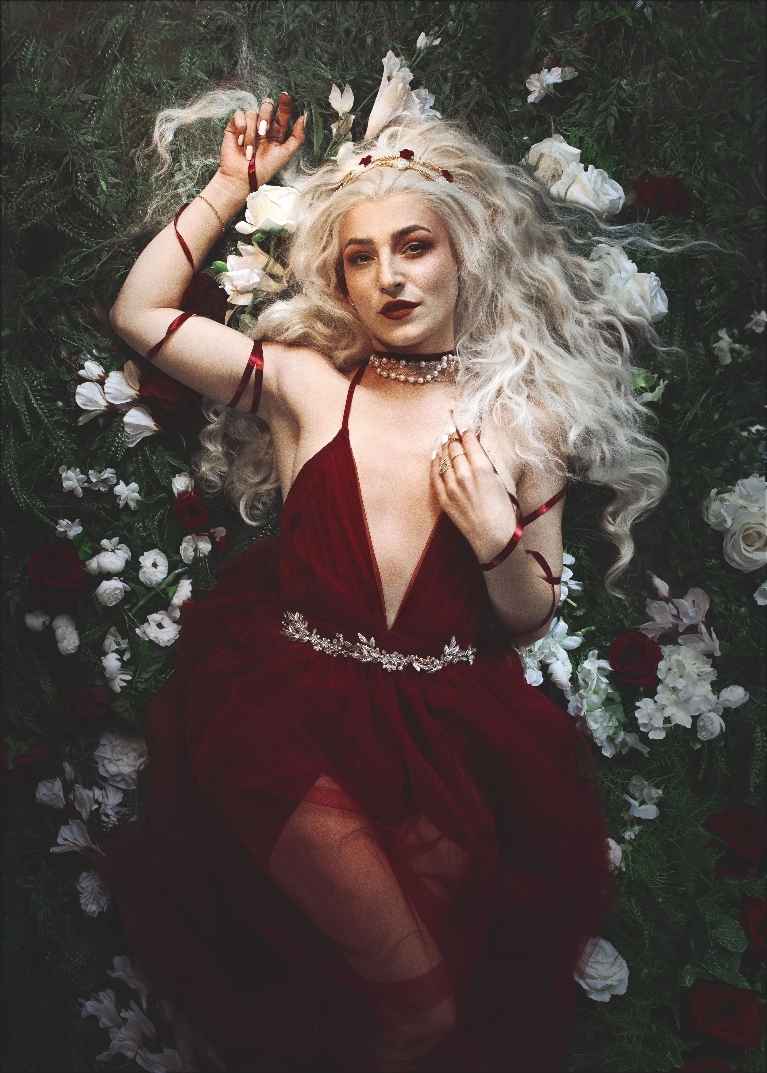 Person with long white hair and a red gown lying on green foliage and flowers.