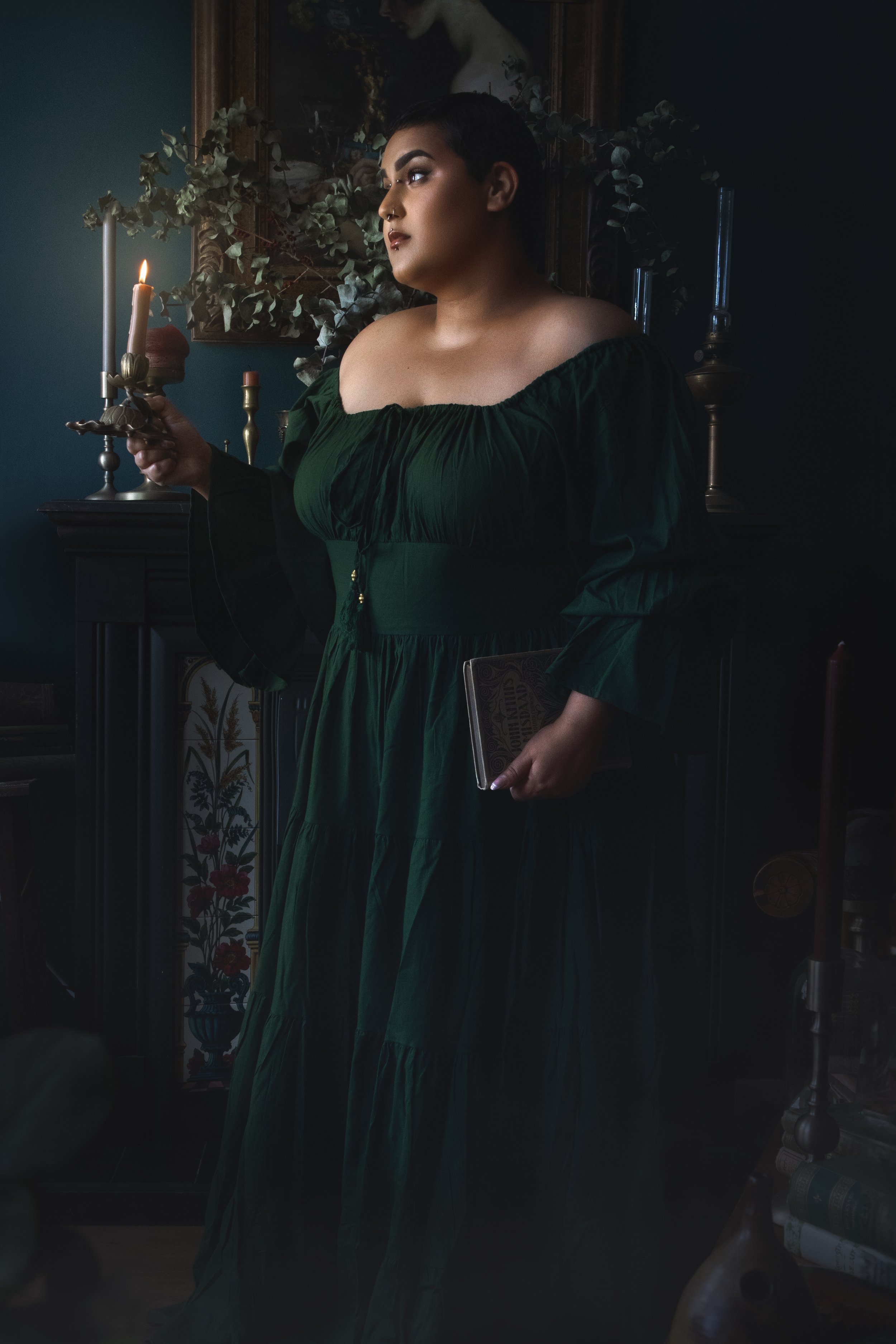 Person in a dark green dress holding a lit candle and a book, standing indoors beside a fireplace with decorative elements, plants, and a painting in the background.