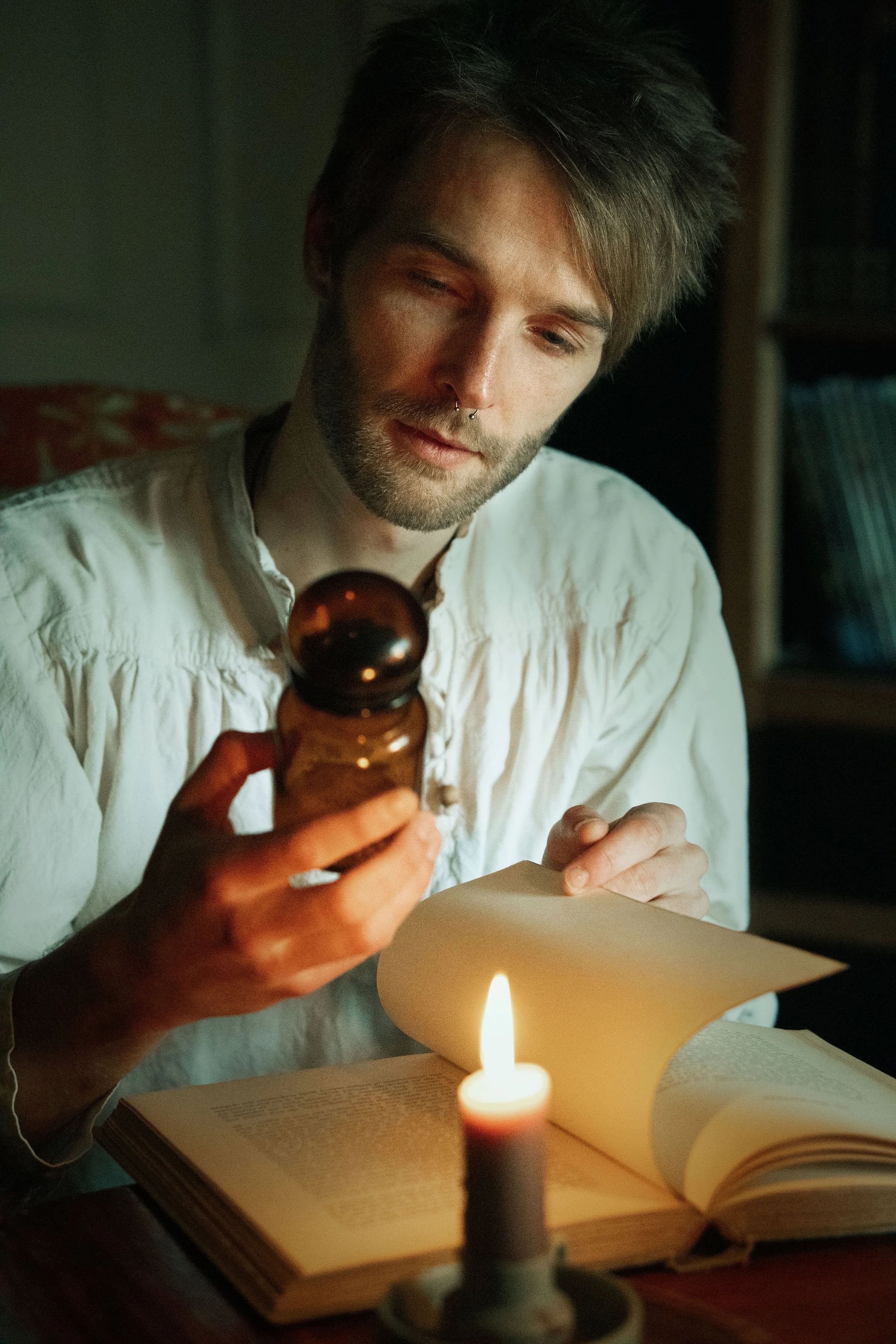 Dreamy fine art portrait of Rico as a scholar reading by candlelight. Painterly fantasy photography inspired by folklore and classical paintings, featuring books, glass bottles and warm light. Moody, romantic storytelling imagery in a fine art studio