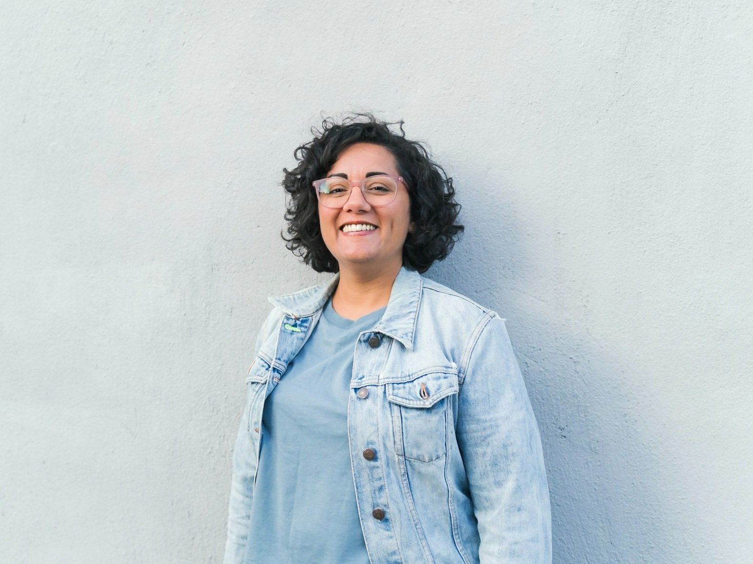 Sumi Fitzgerald, a brown person with dark curly hair and glasses grinning. i'm standing in front of a blank wall.