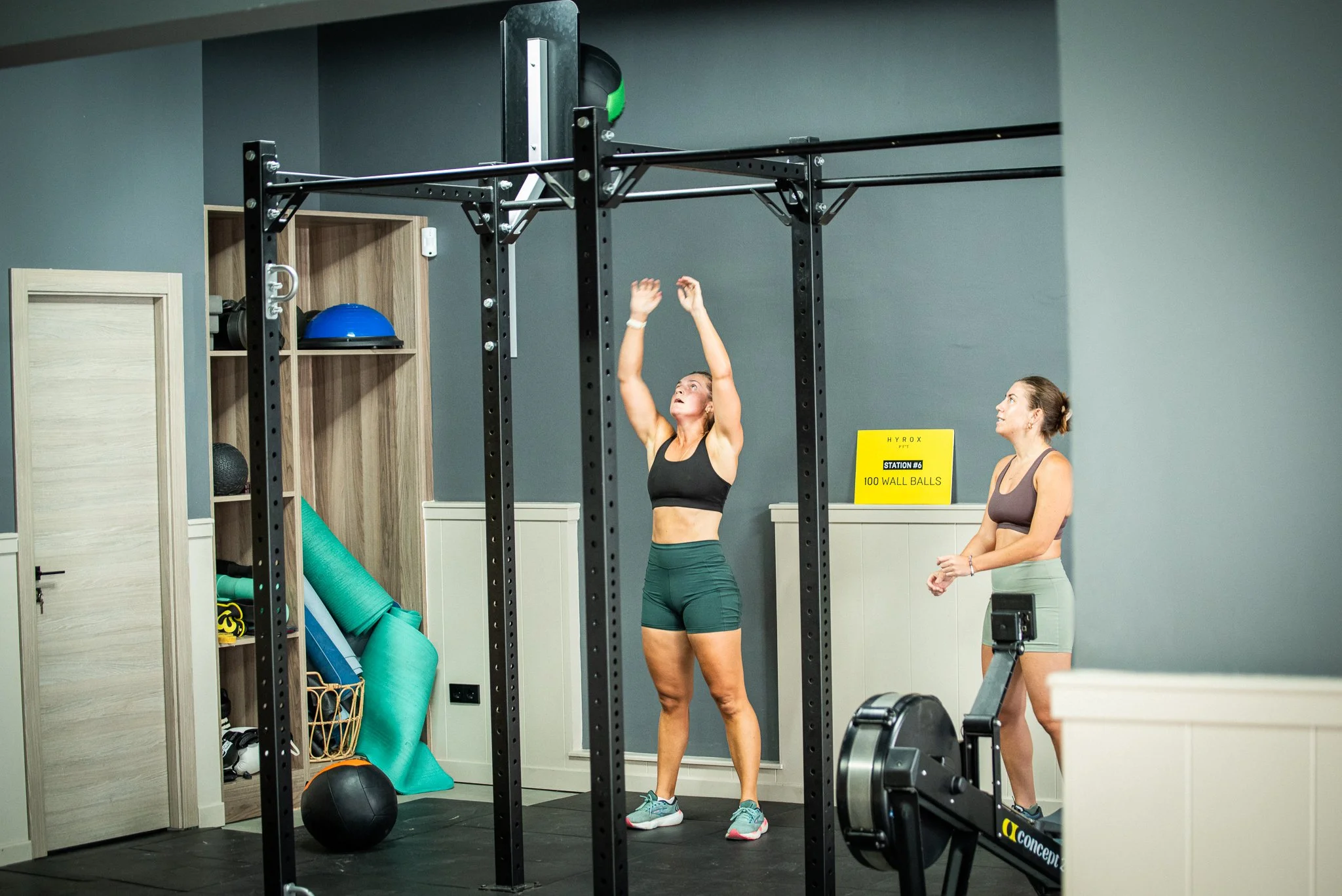 Women in workout clothing in a gym, preparing for exercise, with dumbbells on the floor and a trainer giving instructions.