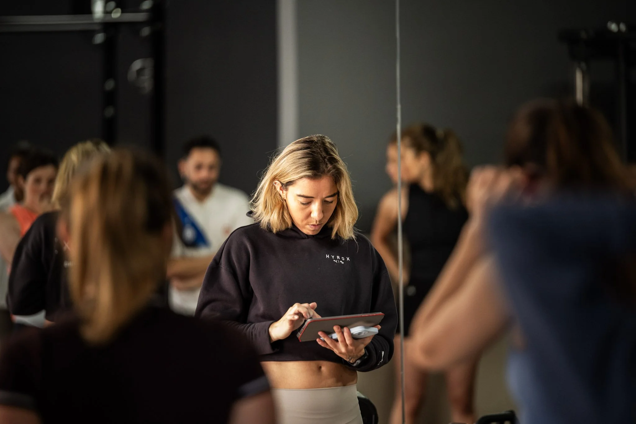 A young woman with blonde hair, wearing a black hoodie, looks at her phone in a dance studio surrounded by other dancers and a mirror.