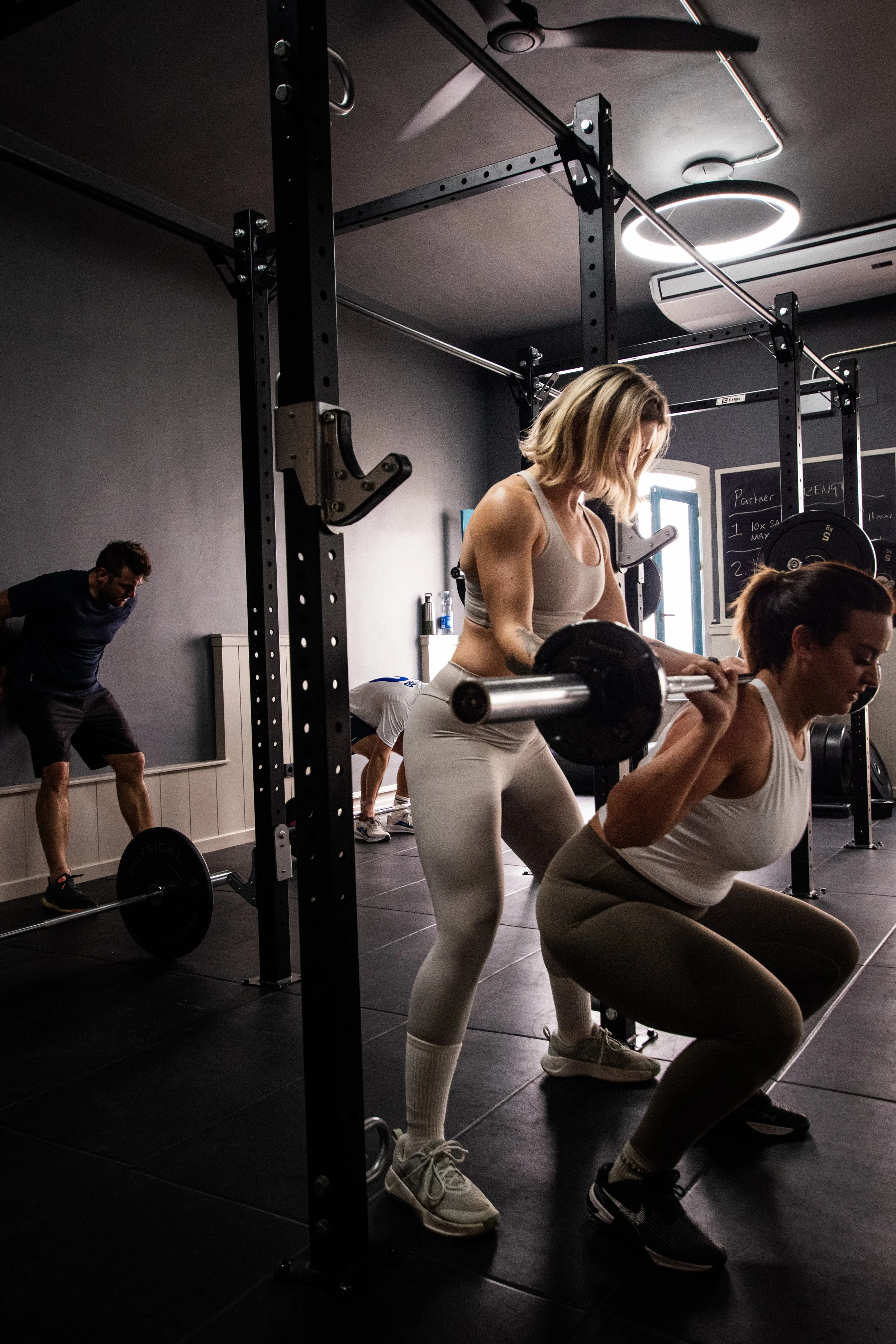 Women exercising with barbells in a gym, with other individuals in the background. The gym has a black interior with fitness equipment.