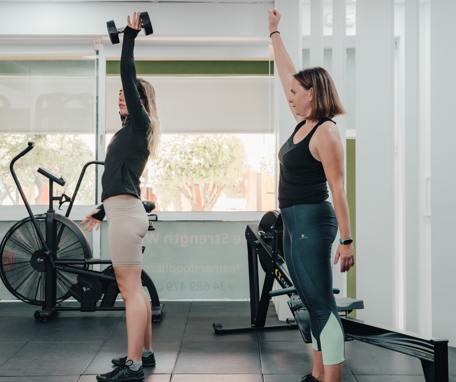 Two women exercising in a gym, with one lifting dumbbells and the other raising her arm in a stretch or pose.