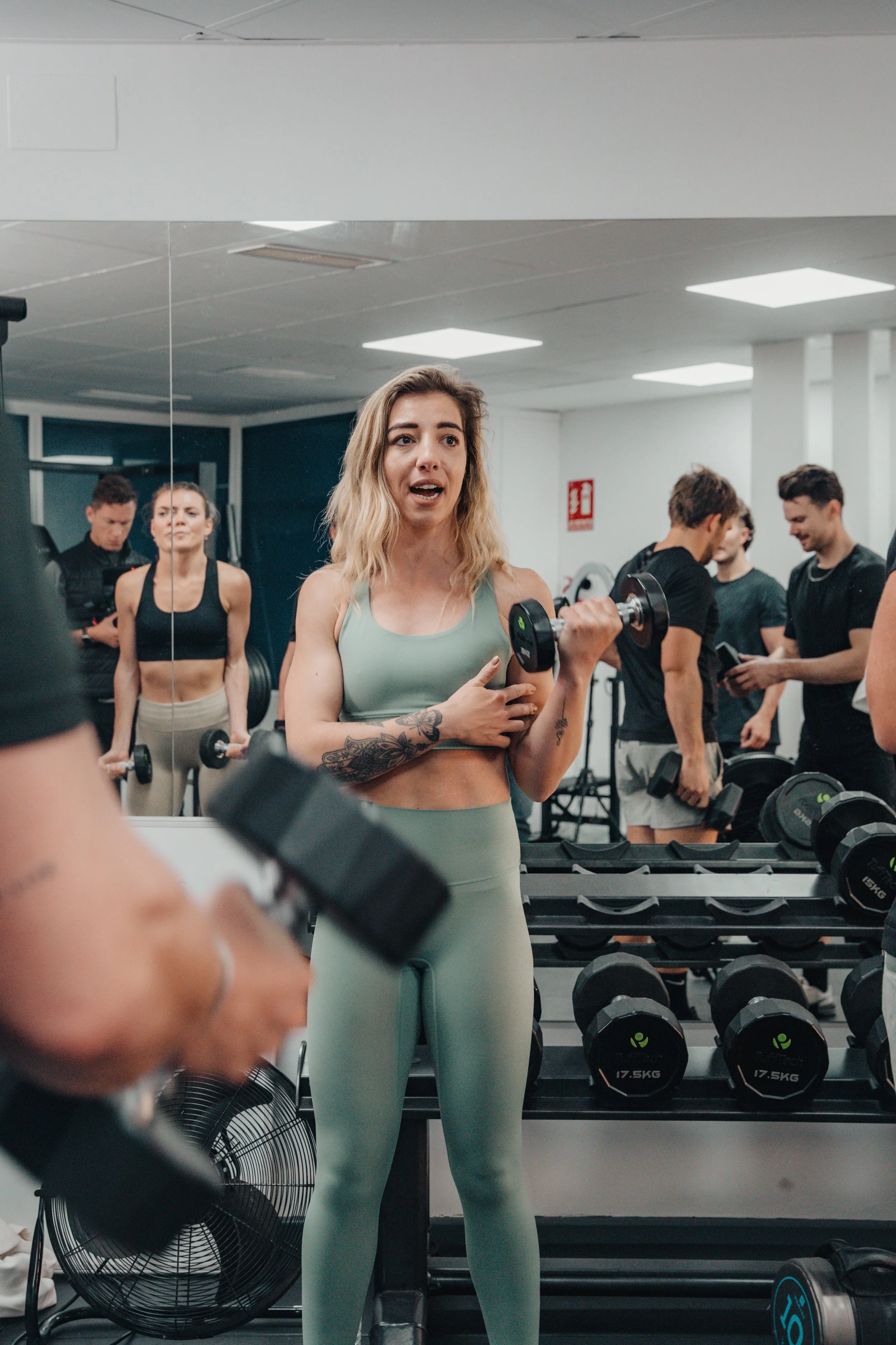 A woman is holding a dumbbell in a gym, surrounded by other people lifting weights and socializing.