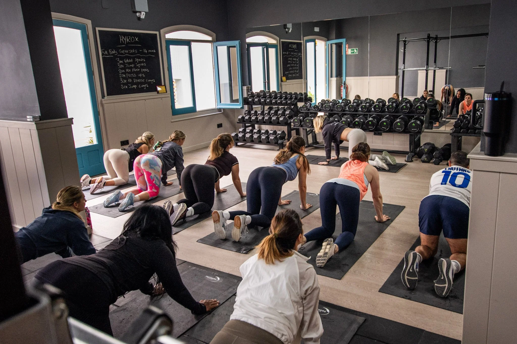 Group of women participating in a fitness class doing on hands and knees exercises on yoga mats in a gym room with mirror wall and dumbbell racks.