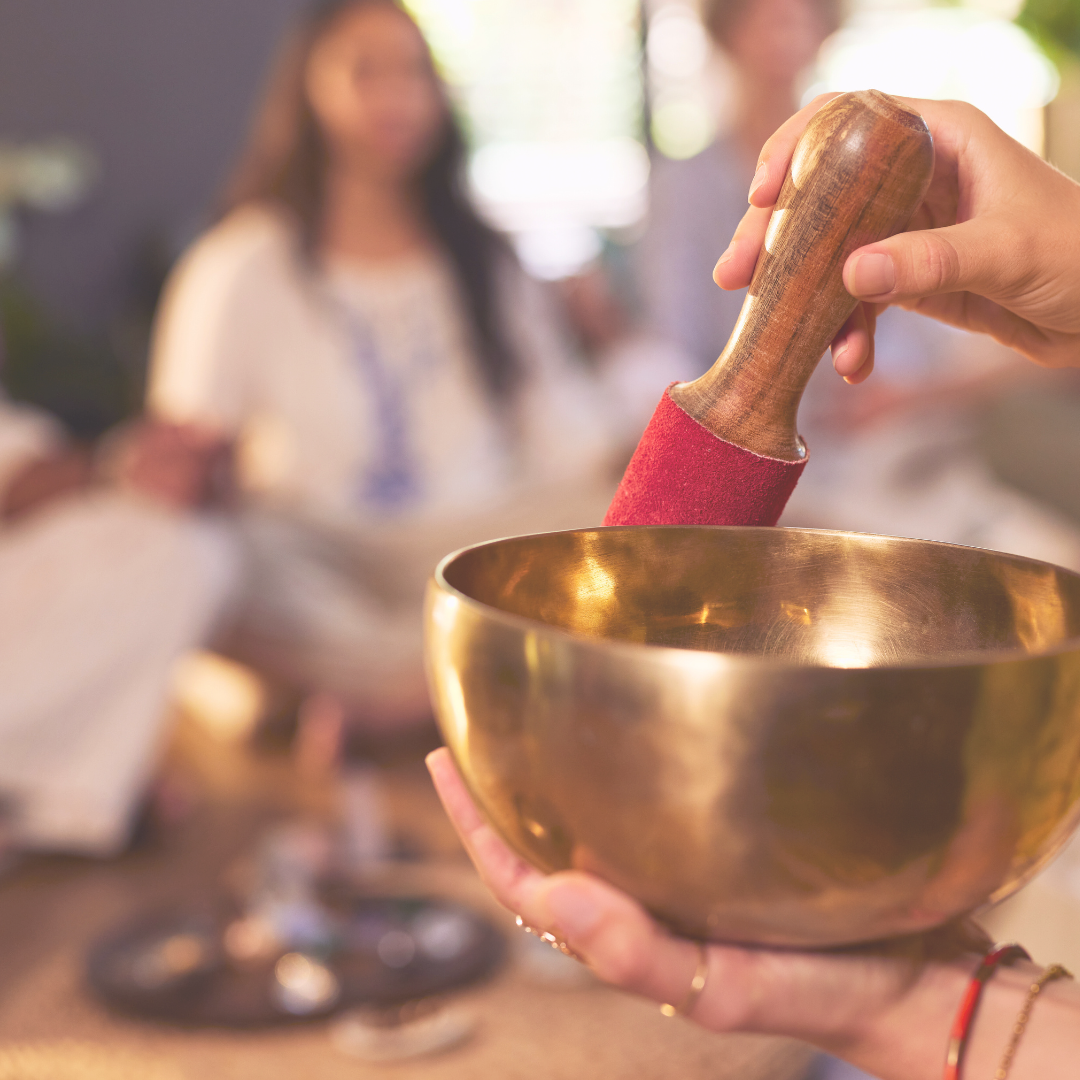 Mano sosteniendo un cuenco de metal dorado y un mazo para tocarlo, en un grupo de personas en un entorno interior con luz natural.