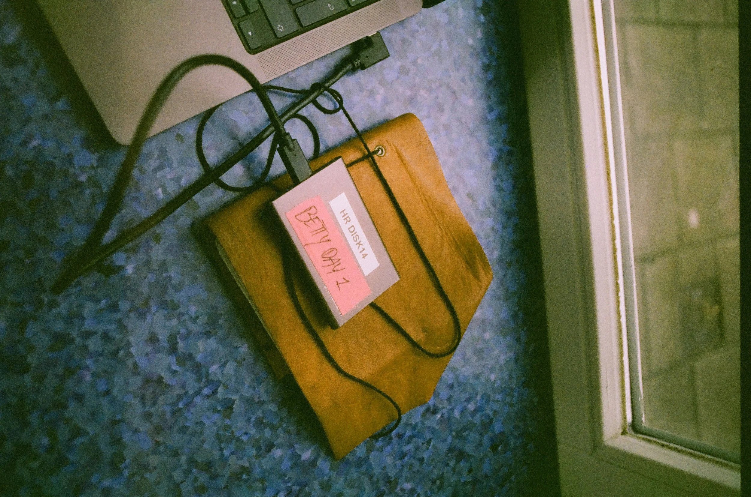 A laptop computer, a notebook with red and white labels, and a telephone cord on a blue speckled countertop near a window.