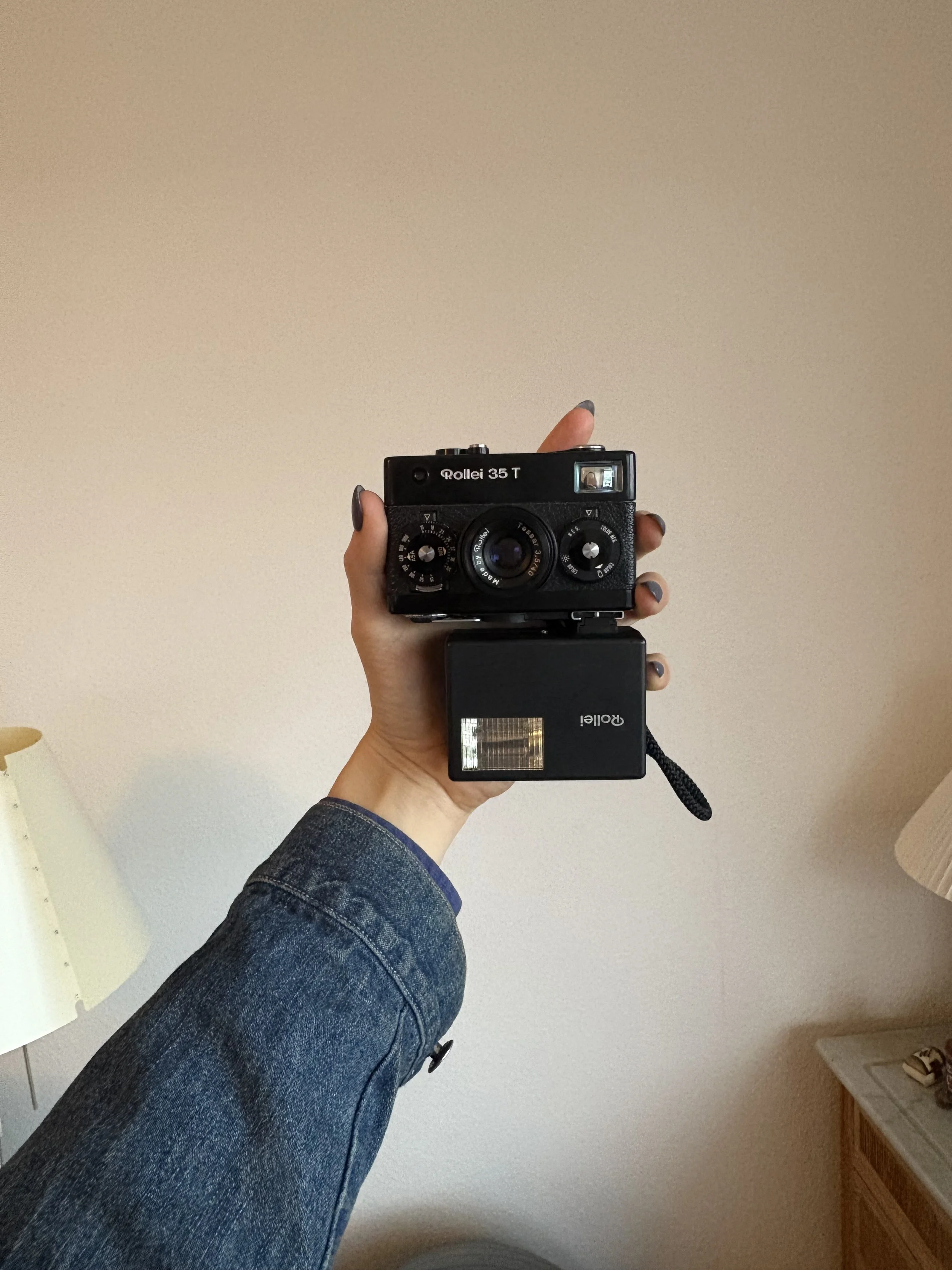 A person holding a vintage black Rollei 35 T camera with a flash attachment, wearing a denim jacket, taking a mirror selfie in a room with beige walls and a wooden cabinet.