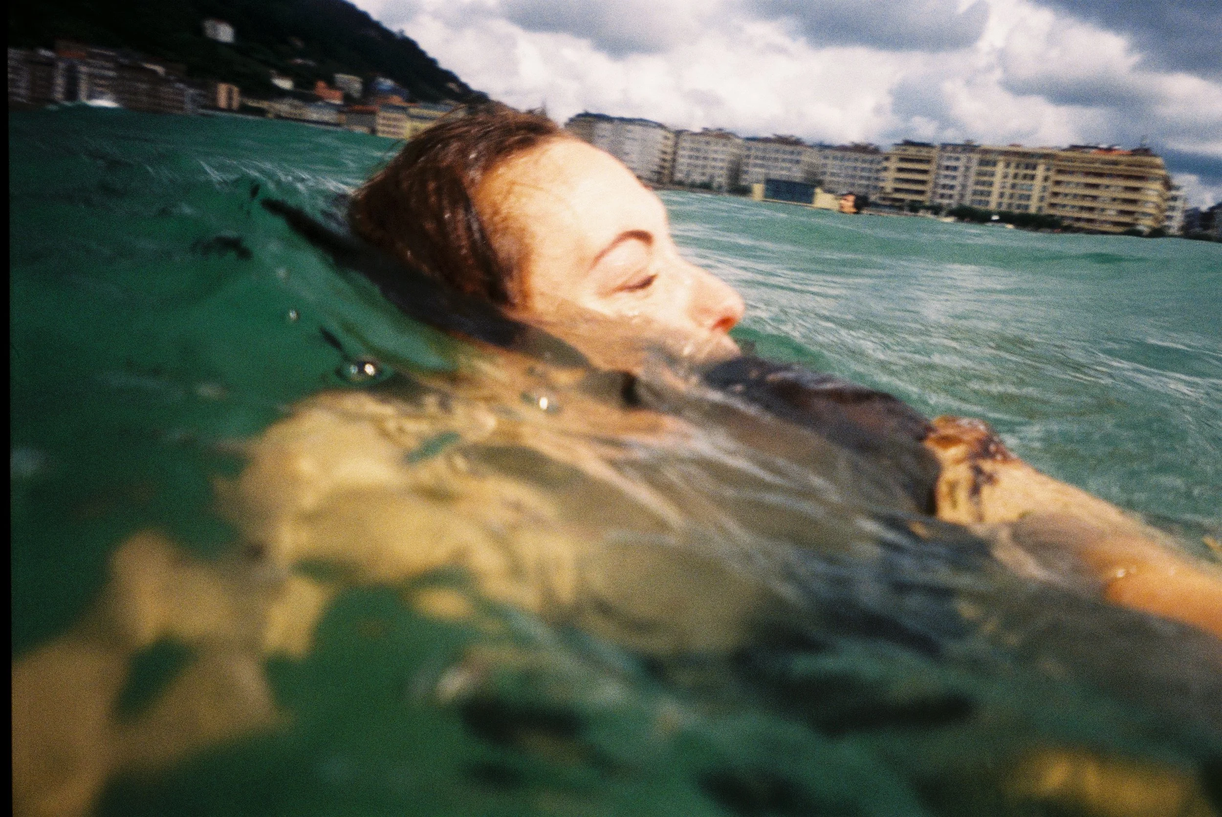 A person with short hair floating on a green body of water with their eyes closed, against a backdrop of a city skyline and cloudy sky.