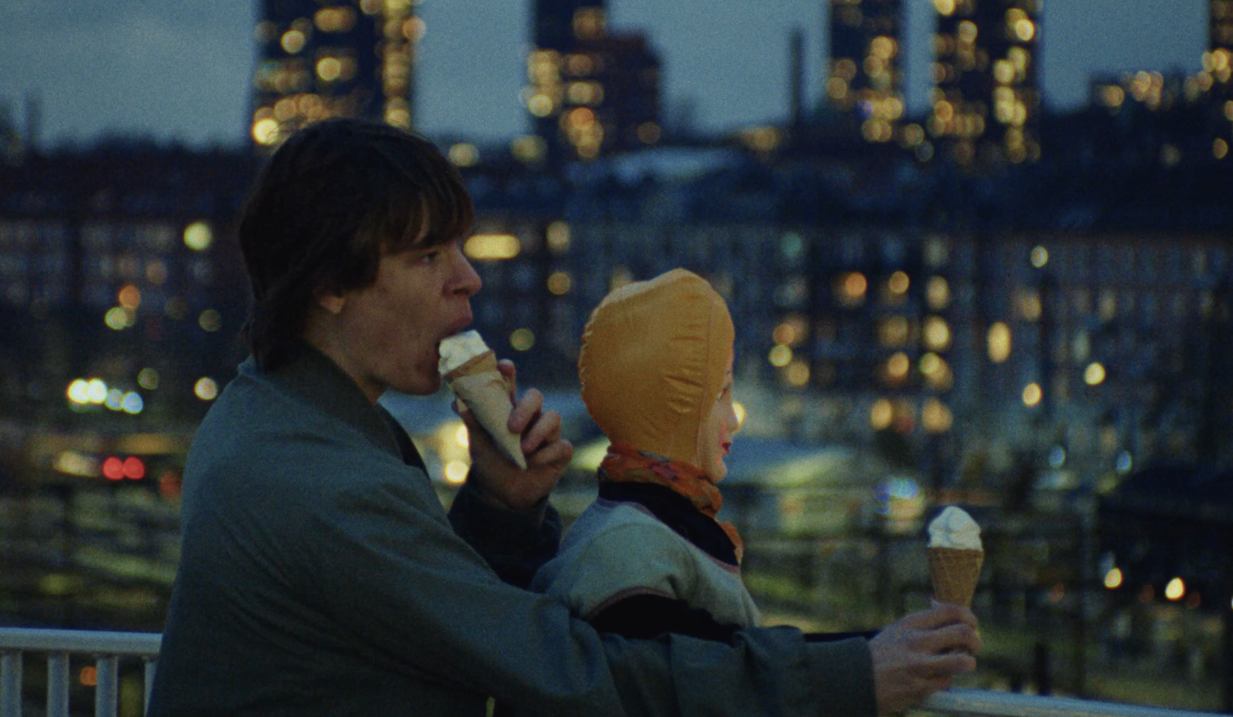 A young man with dark hair is eating ice cream cone while sitting on a railing at dusk. A young woman with a yellow hood and colorful scarf is also eating ice cream beside him. The city lights are blurred in the background.