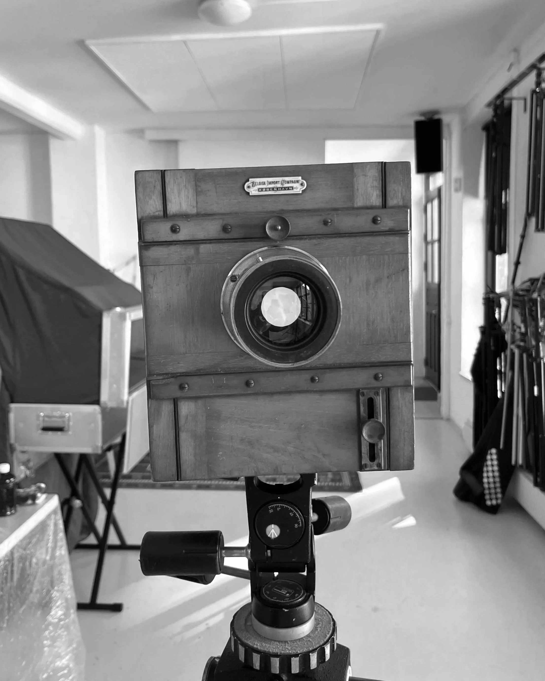 A vintage large format camera mounted on a tripod in a room, with a perspective looking through the camera lens toward a white wall.