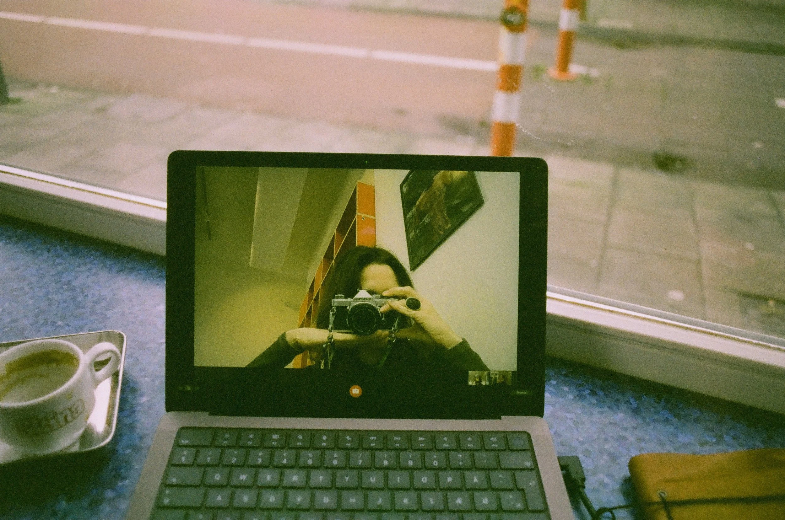 Reflection of a woman taking a photo with a camera in a laptop screen on a cluttered desk, with a cup of coffee on the left and a yellow object on the right, in a room with a window and blinds.