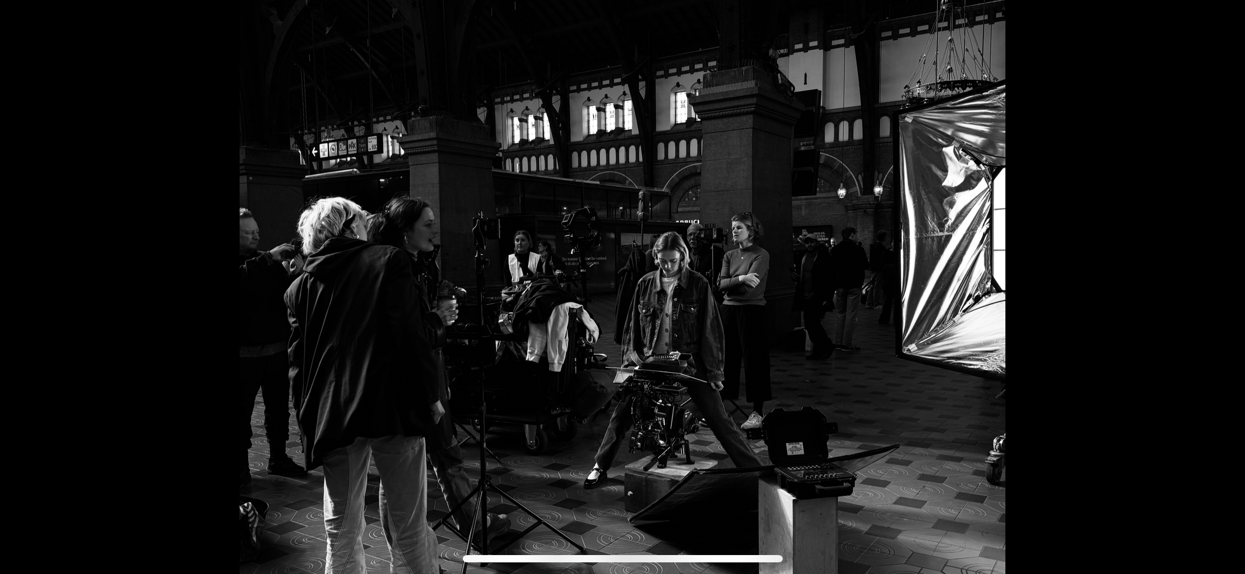 A black and white photo of a film or photography crew working inside a large, historic train station with high arched windows and ornate architecture. Several people are gathered around a camera and lighting equipment on a temporary set, preparing fo