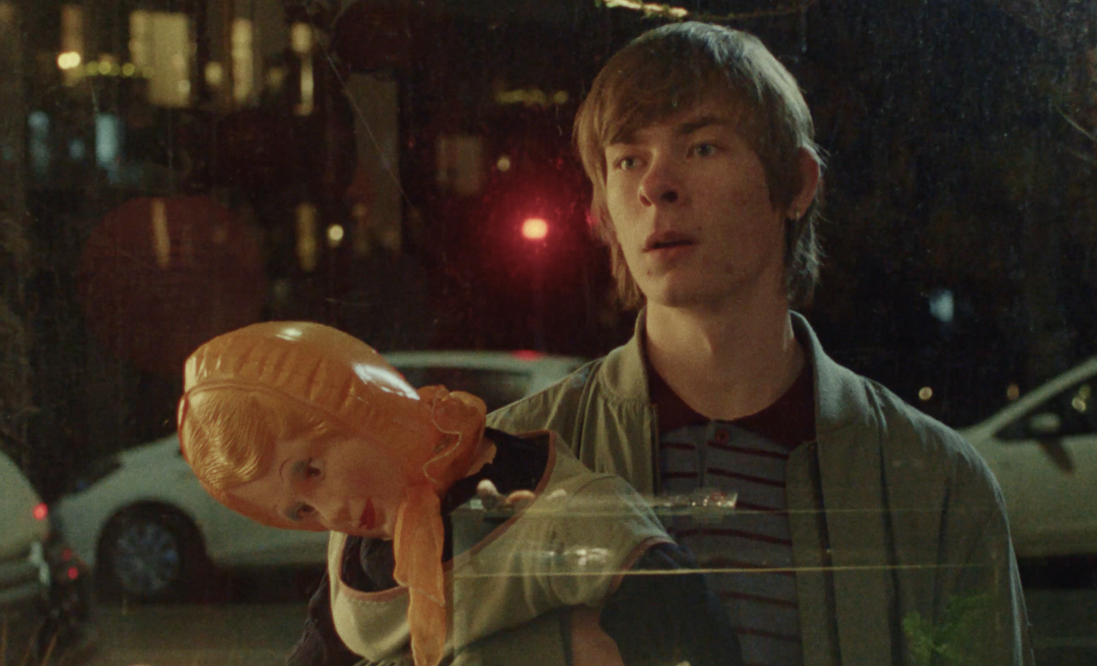 A young man with light brown hair standing outside at night, looking at something off-camera. He is wearing a tan jacket over a striped shirt and holding a bag of something orange with a biscuit shape.