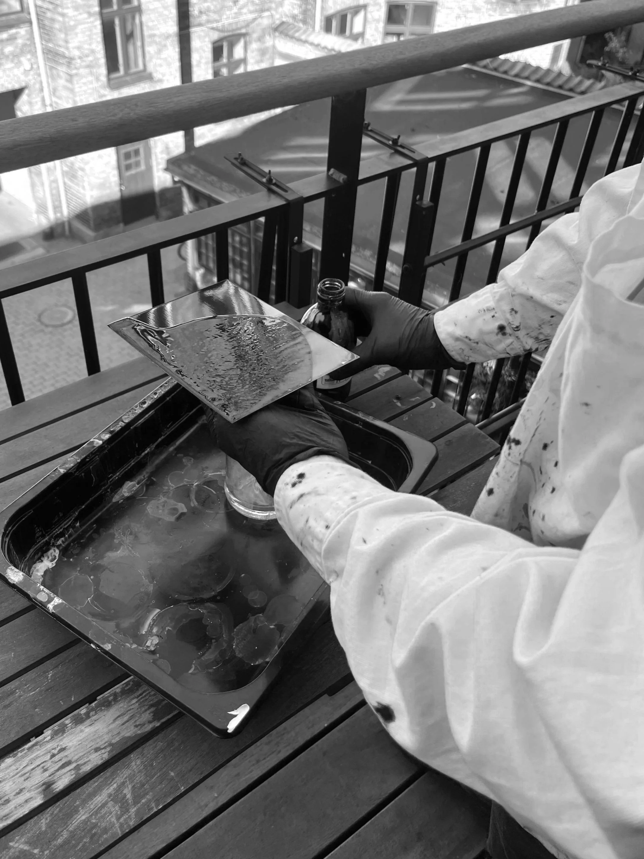 A person in a white smock and gloves holding a small bottle and a metal tray with a liquid substance, sitting on a wooden bench near a metal railing on a balcony with city buildings in the background.