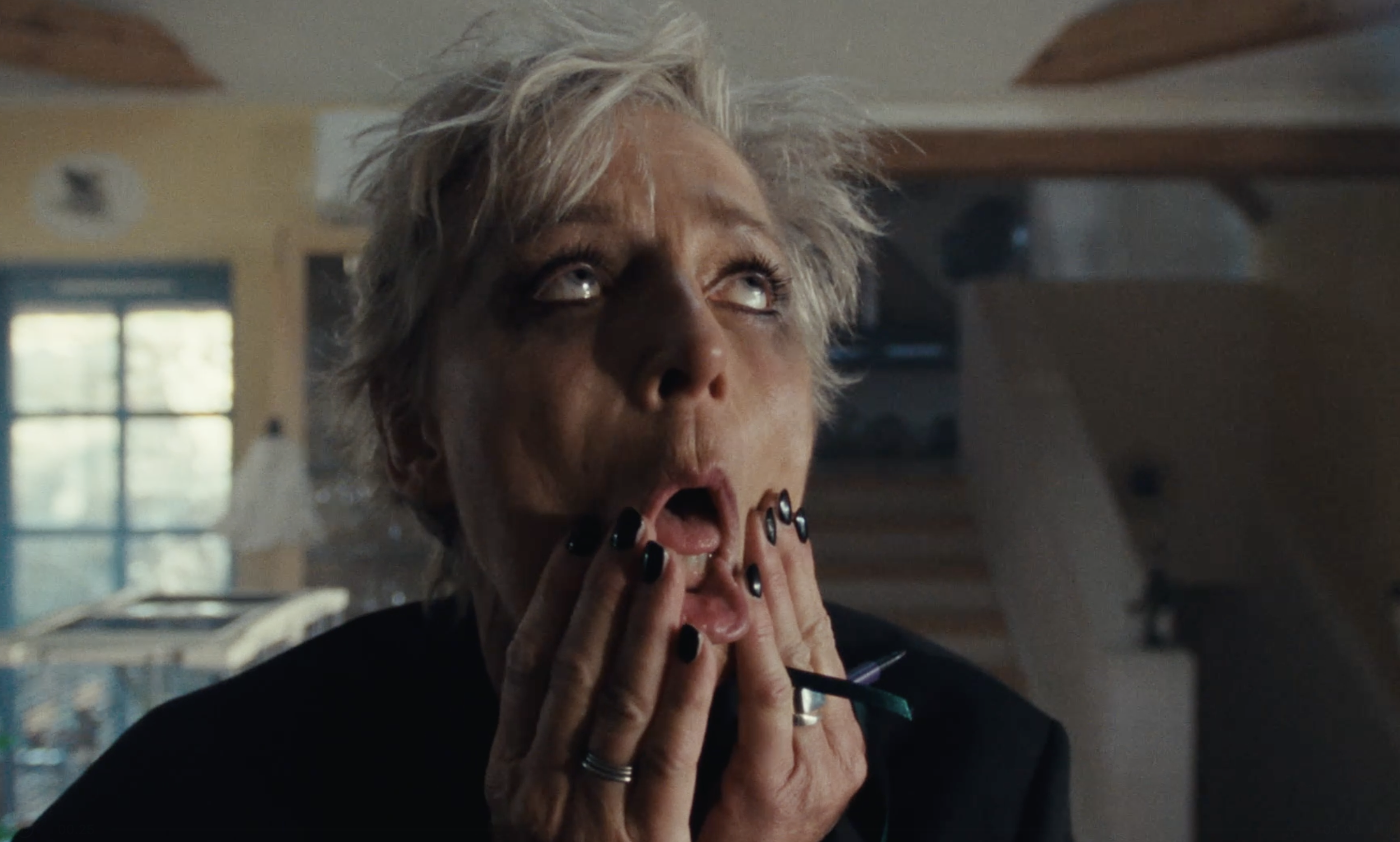 A woman with short, messy blonde hair and black nails appears surprised, with her hands on her face, in a kitchen with a window and cabinets in the background.