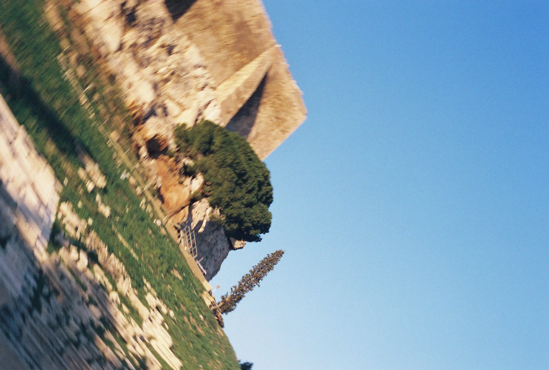 The image features the Great Pyramid of Giza against a clear blue sky, surrounded by trees and rocky terrain, with some greenery and a stone wall in the foreground.