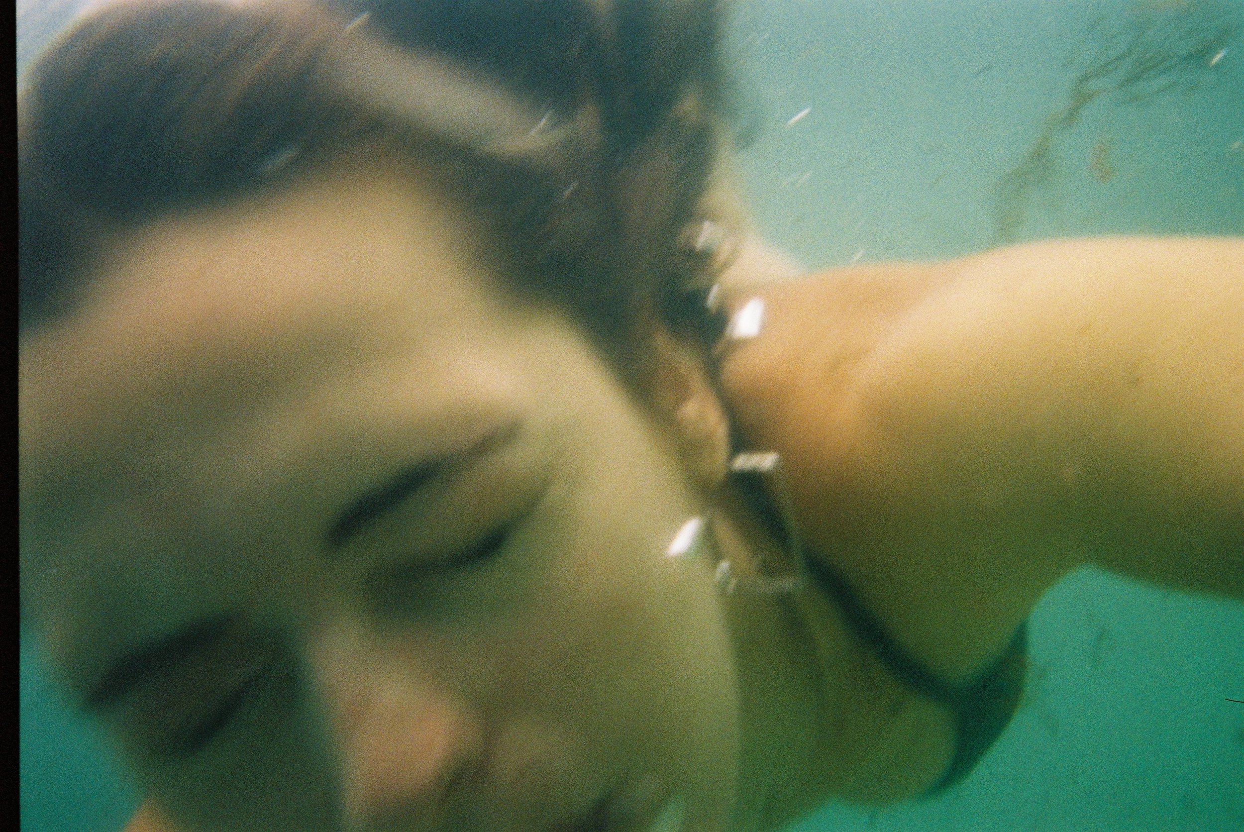 A close-up, underwater shot of a woman with long dark hair, wearing a black swimsuit, holding a lipstick.