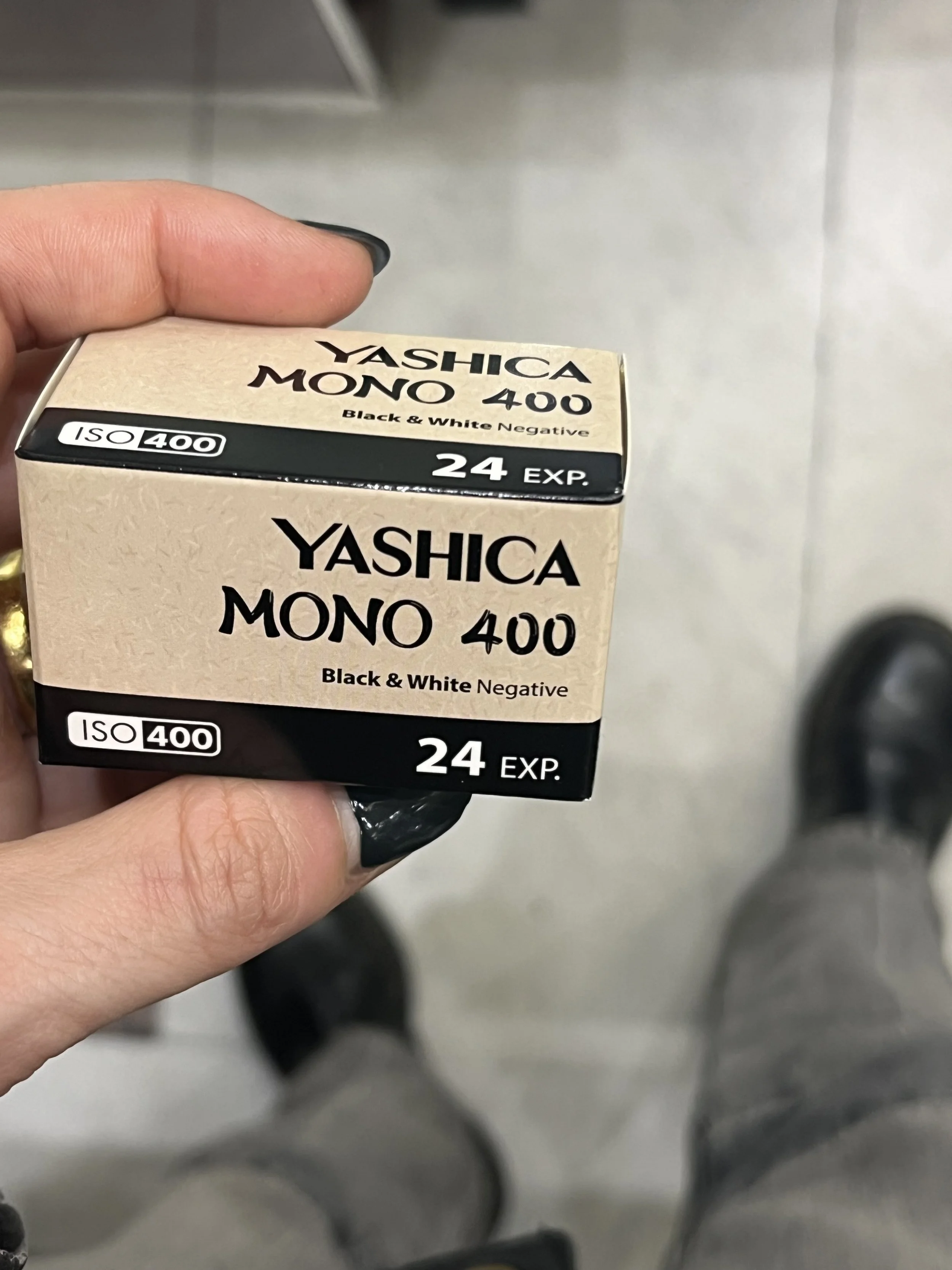 Close-up of a person's hand holding a box of Yashica Mono 400 film with a gray tiled wall background.
