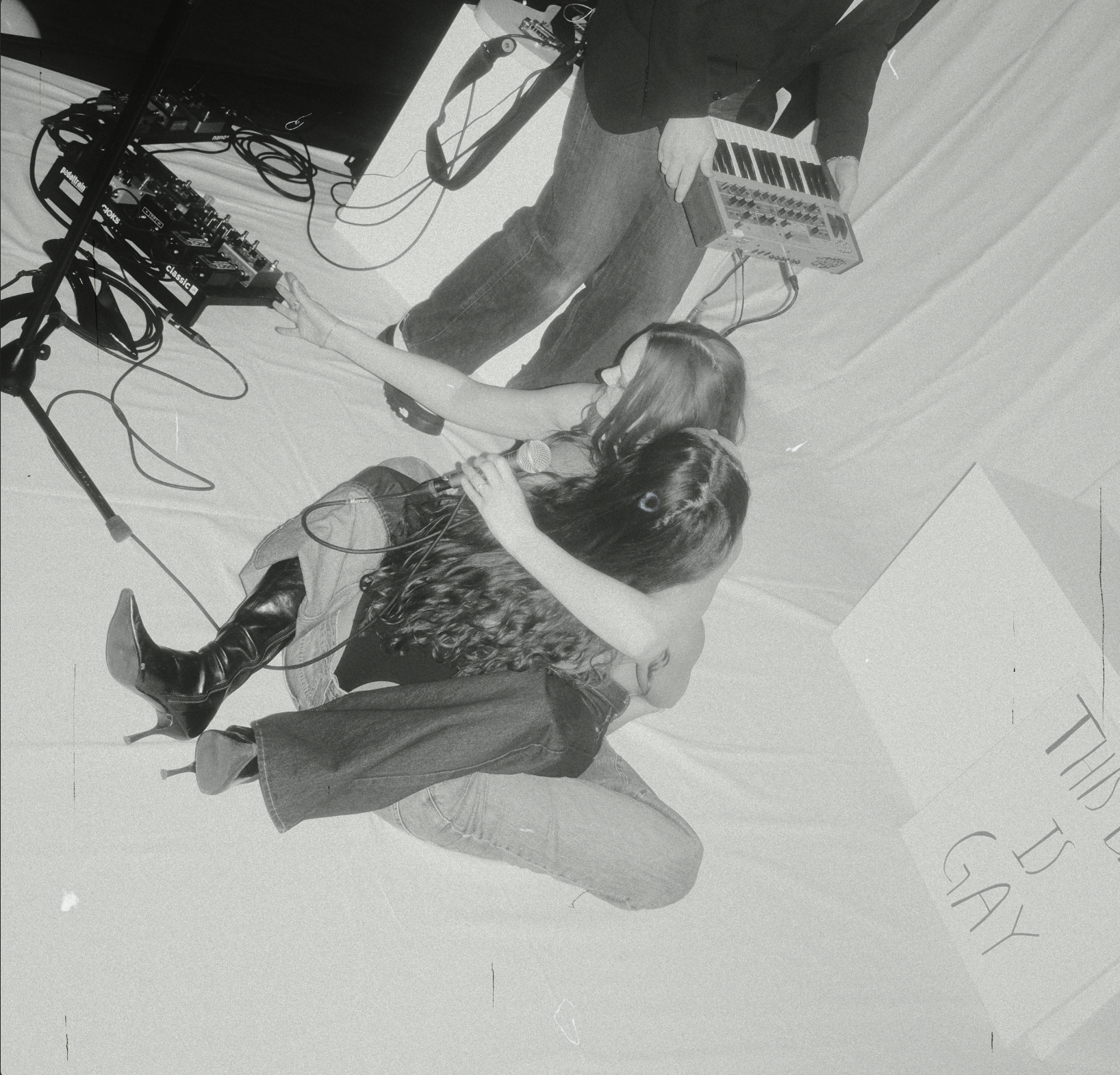A woman with long hair wearing boots and a jacket is sitting on the floor while a person with a long-sleeved shirt and jeans stands beside her playing a synthesizer. There is electronic music equipment on a table nearby, including a mixer and cables.