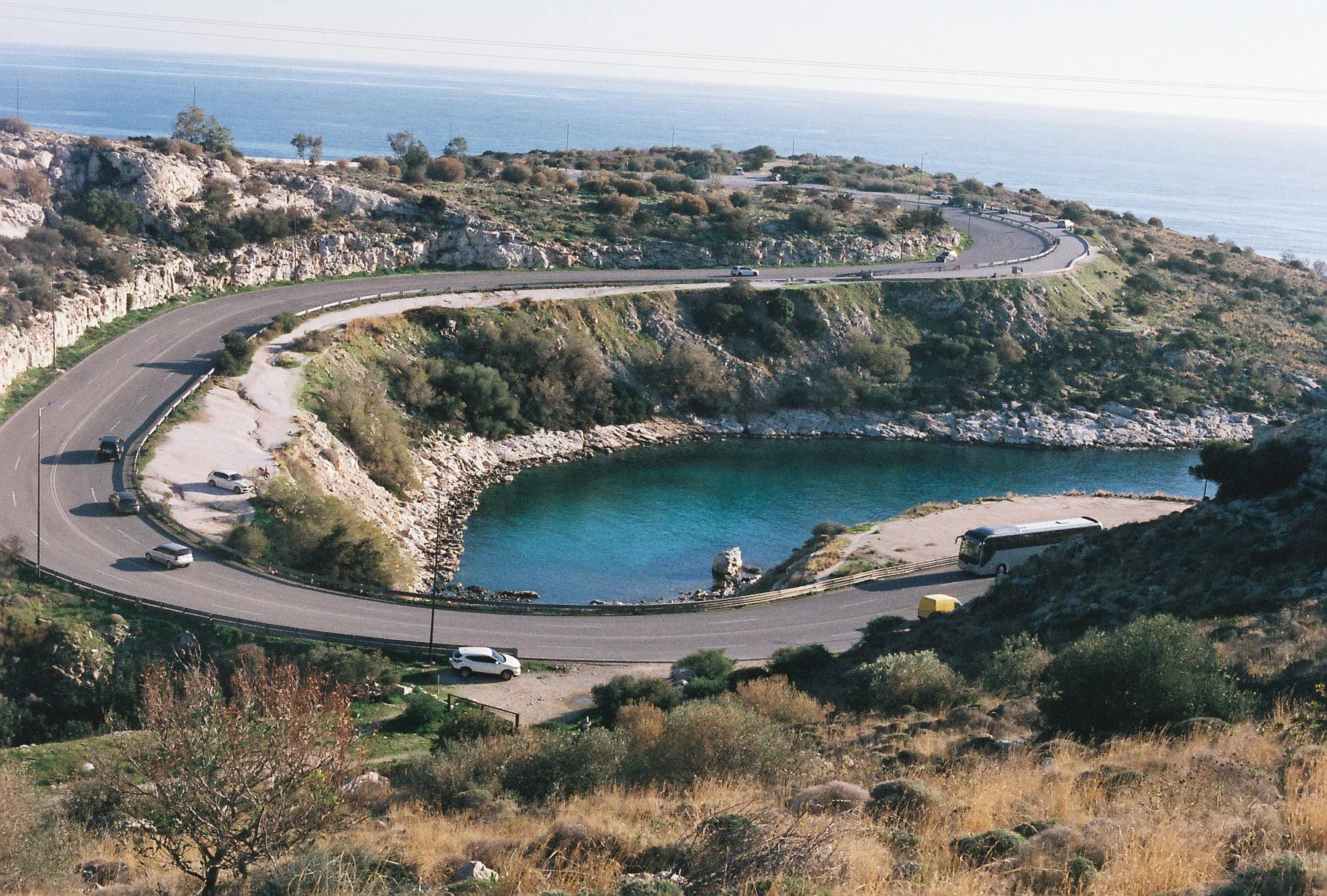 A winding coastal road with cars and a bus, overlooking a body of water and rocky cliffs with sparse vegetation and trees.