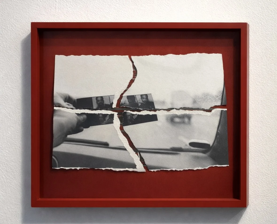 Mixed media artwork with a photograph of a torn mirror and a hand holding a small photo strip, mounted in a red frame on a white wall.