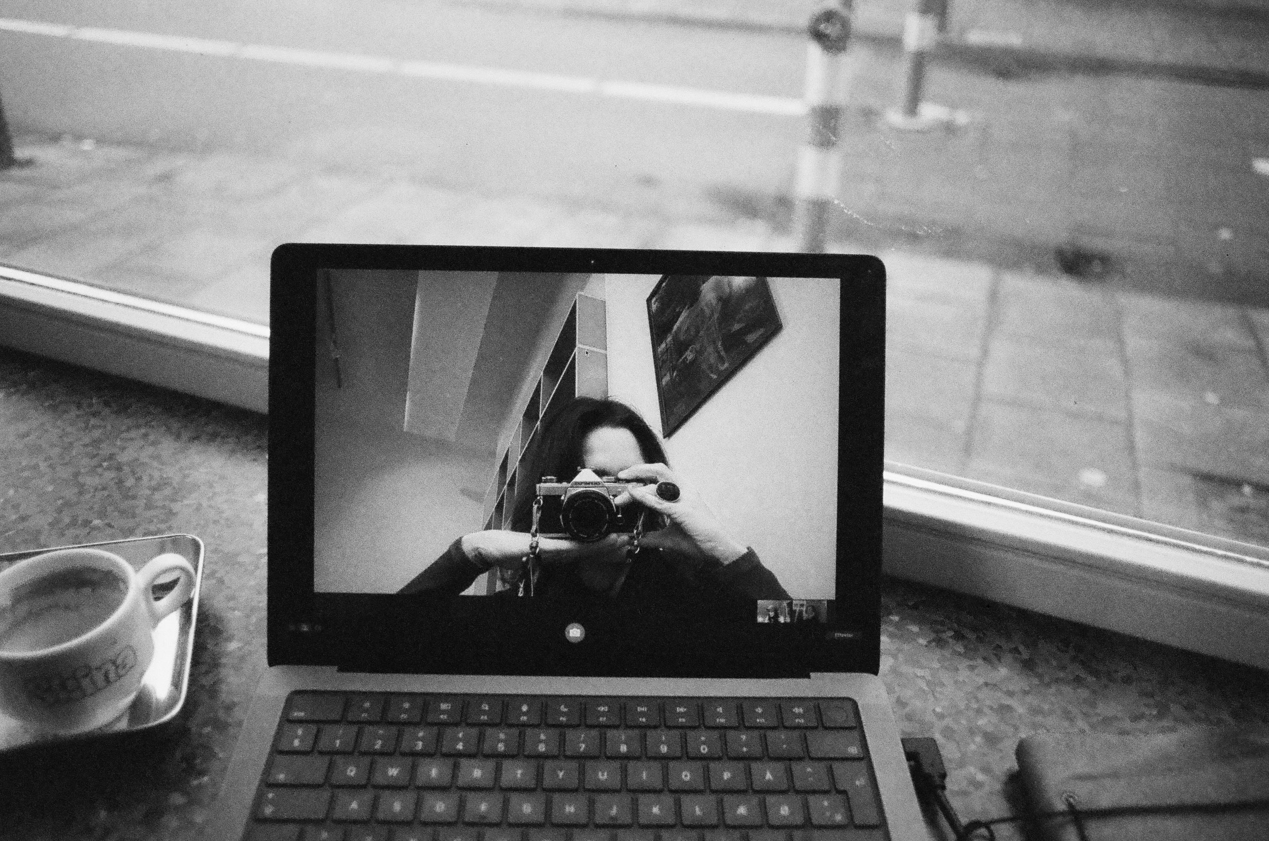 A person taking a photo of their reflection in a computer monitor, with a keyboard and two coffee cups on a table nearby, in a black-and-white photo.