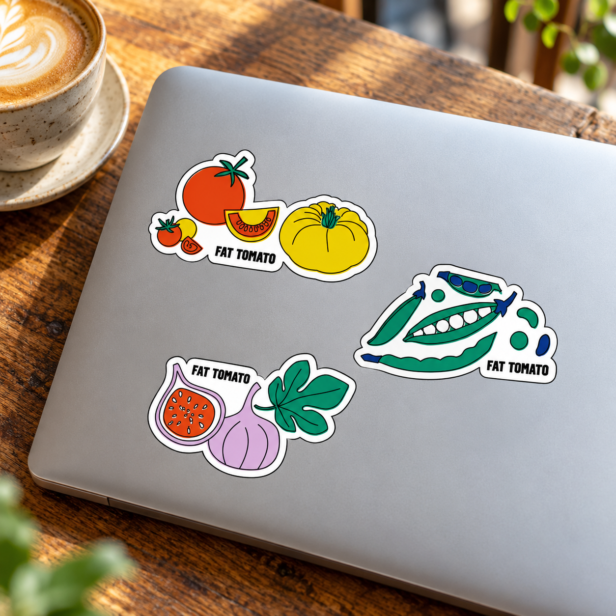 Tomato, peas and beans, and fig stickers on a silver laptop beside a cup of coffee.