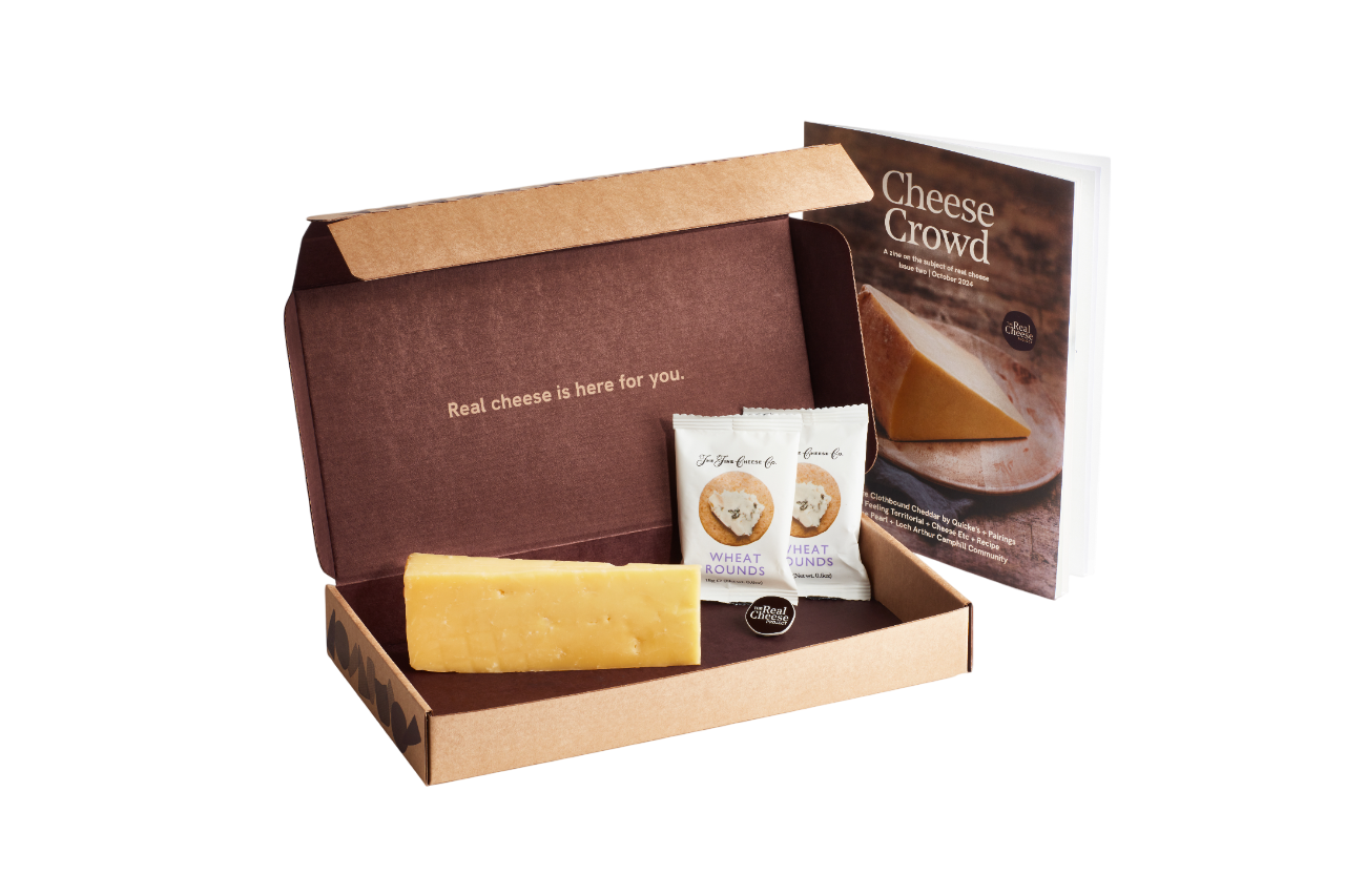 The image shows a cheese tasting kit with a block of cheese, two packets of wheat rounds, a informational booklet, and a small label, all inside a brown box with the phrase 'Real cheese is here for you.' printed on the inside.