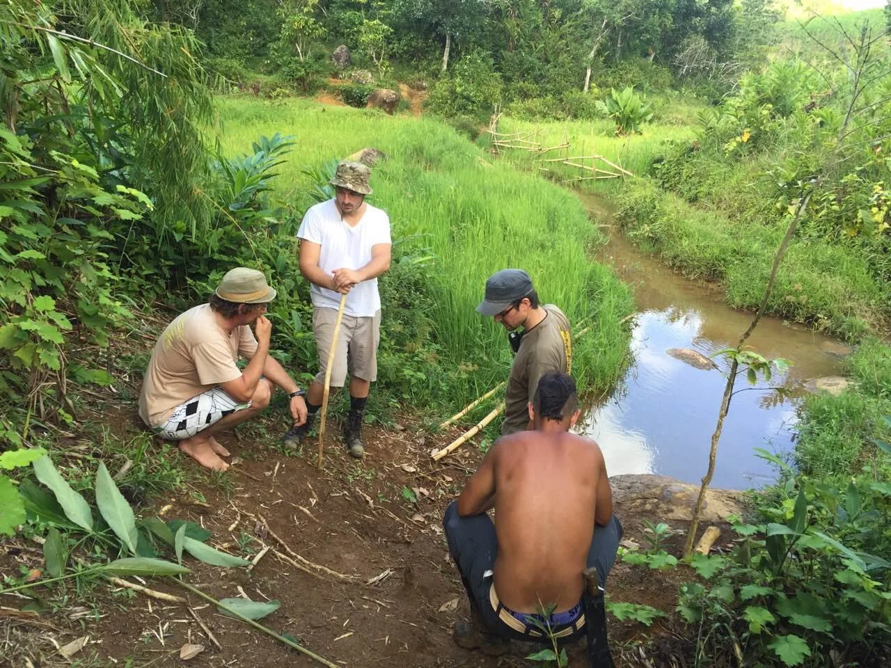 Cinq hommes, dont un nu, près d'une rivière dans une zone verte avec de la végétation dense, discutent devant un terrain en terre.