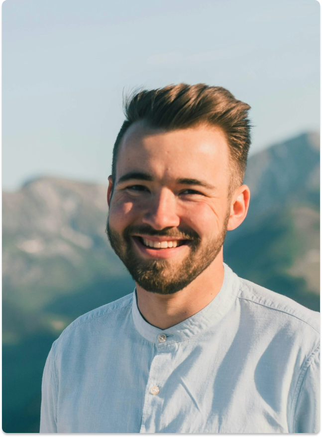 Smiling man with short, styled brown hair and a beard, wearing a light-colored button-up shirt, outdoors with mountains and clear sky in the background.