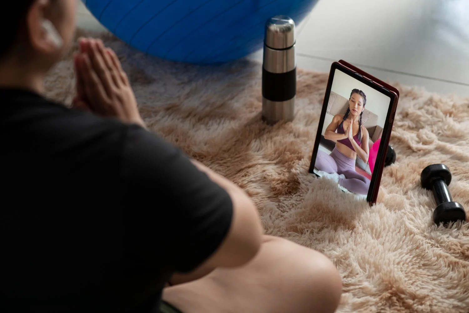 A woman practicing yoga at home, seen through a video call on a tablet. She is sitting cross-legged with hands in a prayer position. The setting includes a fluffy rug, dumbbells, a water bottle, and a large exercise ball.