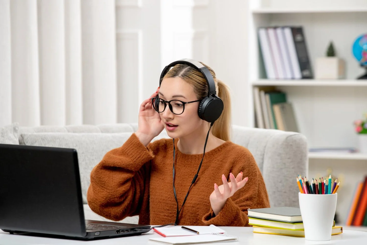 Young woman with glasses and a brown sweater, wearing headphones, sitting at a table with a laptop, notebooks, and a pencil holder with colorful pens, in a bright room with bookshelves.