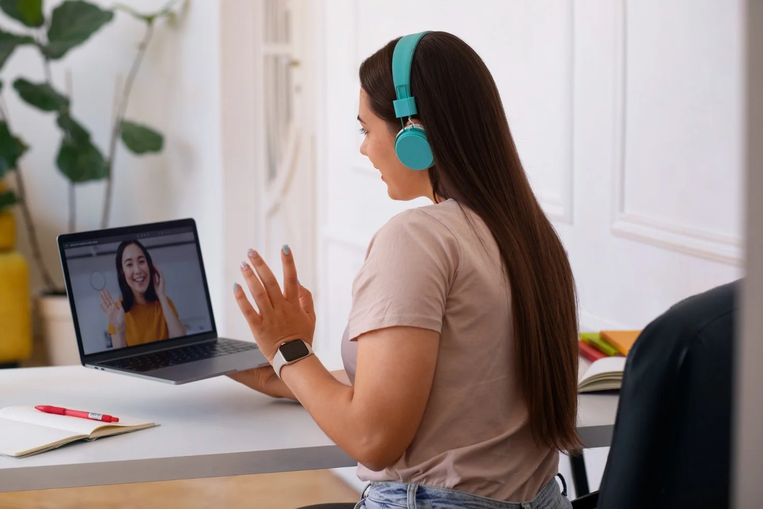 A woman with long brown hair and a smartwatch on her wrist is sitting at a desk, wearing teal headphones, and engaging in a video call on her laptop with a smiling woman waving.