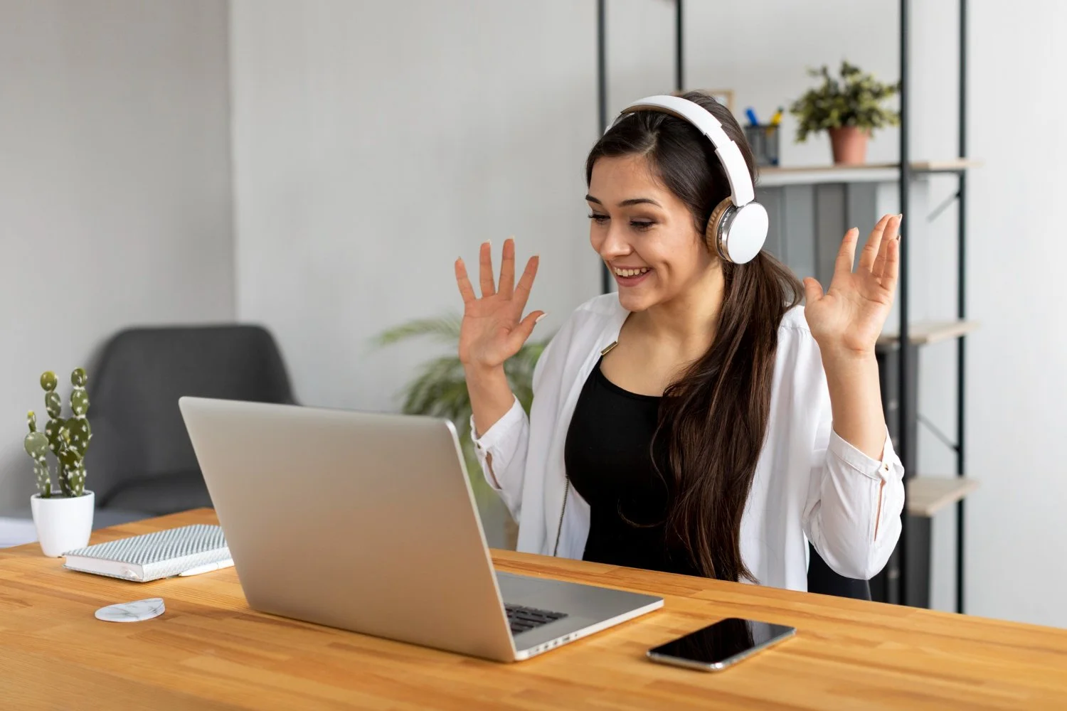 A woman with long brown hair wearing headphones smiling and raising her hands in a video call or virtual meeting at a wooden desk with a laptop, smartphone, and notebook in a modern office.