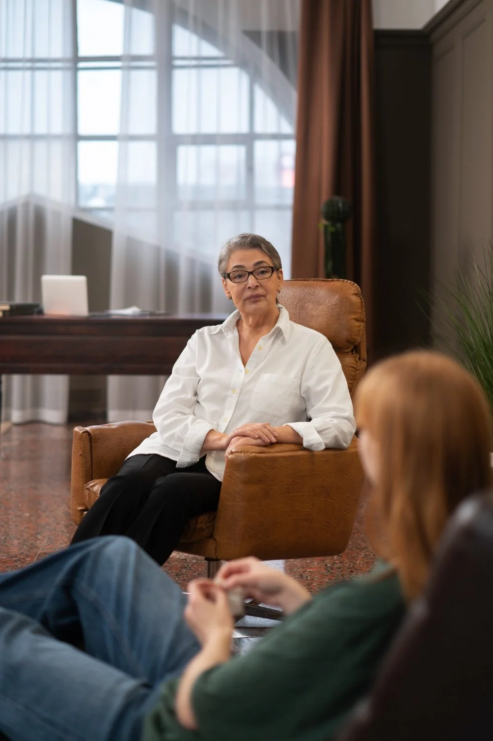 An elderly woman with glasses wearing a white shirt sitting in a brown chair, speaking to a person with long red hair sitting in front of her in an office setting with large windows and brown curtains.