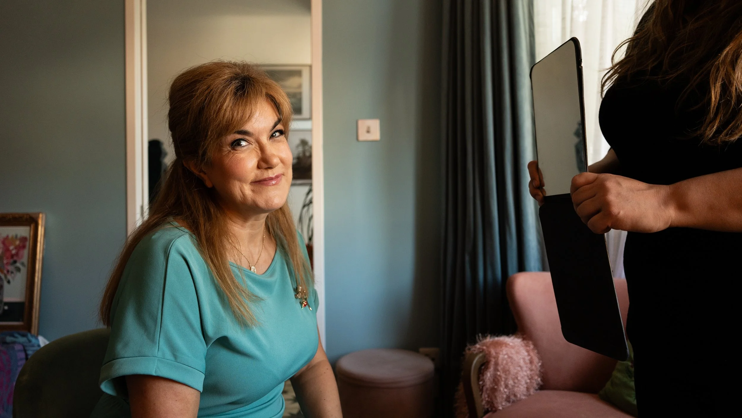 A woman with light brown hair and a blue top sitting indoors, looking at the camera with a slight smile, while another person holding a tablet is partially visible on the right.