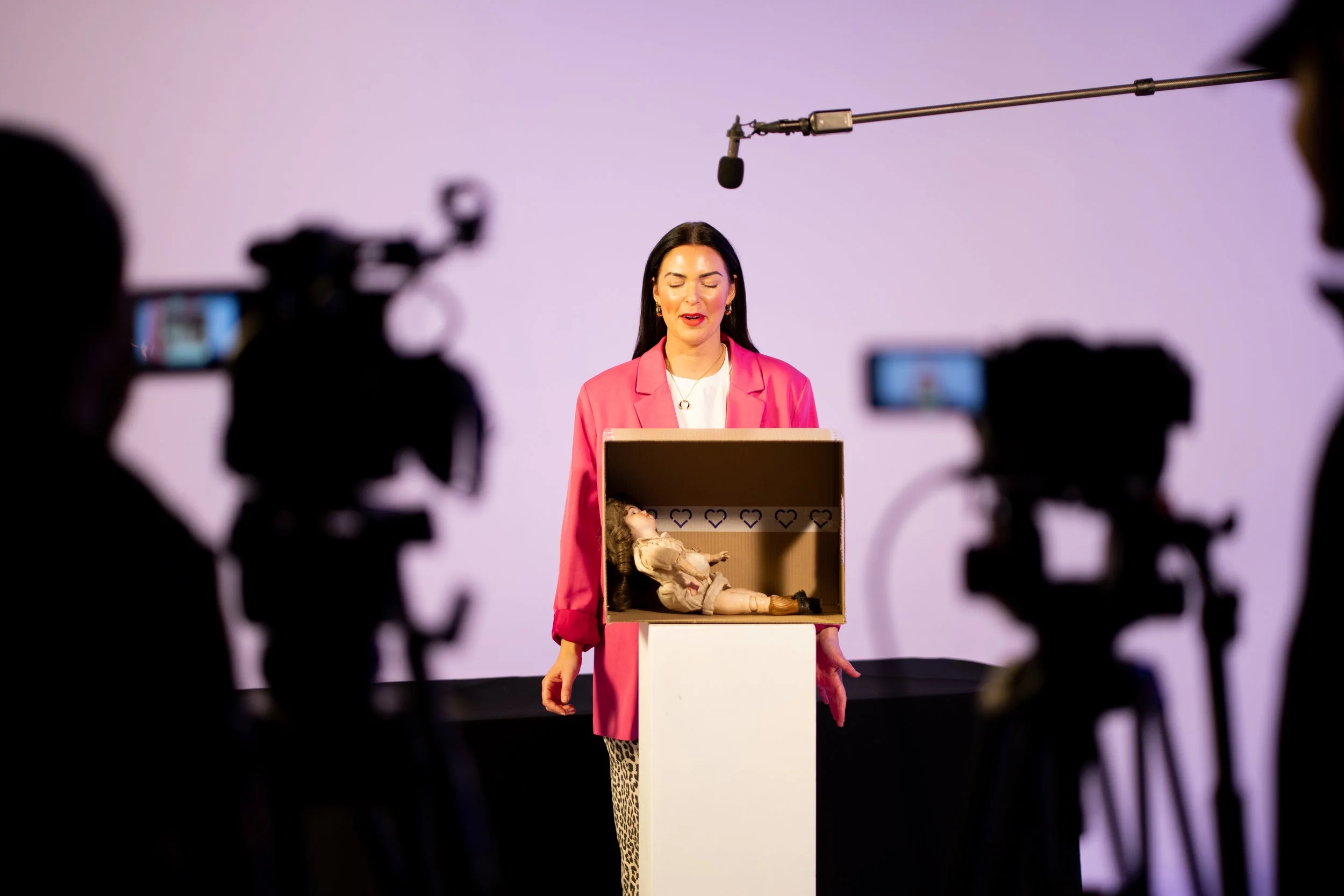 A woman in a pink blazer stands behind a podium with a box on it, inside of which is a doll dressed in beige. She is in front of a purple background, surrounded by cameras and recording equipment, with a microphone overhead.