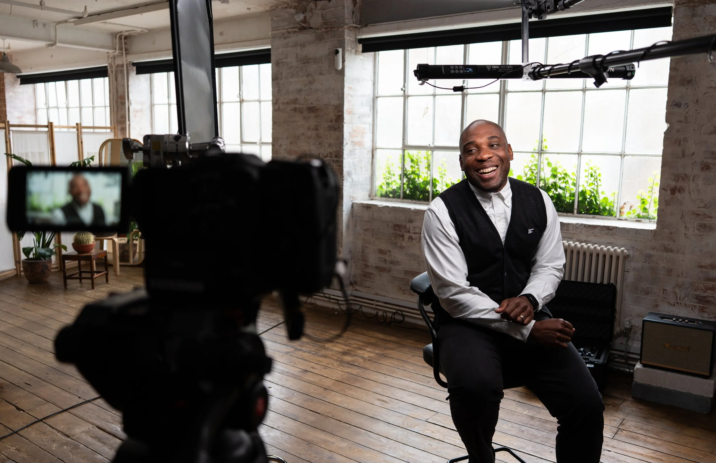 A man smiling during a video recording in a bright, industrial-style room with large windows, wooden floors, and decor including plants and a Marshall amplifier.