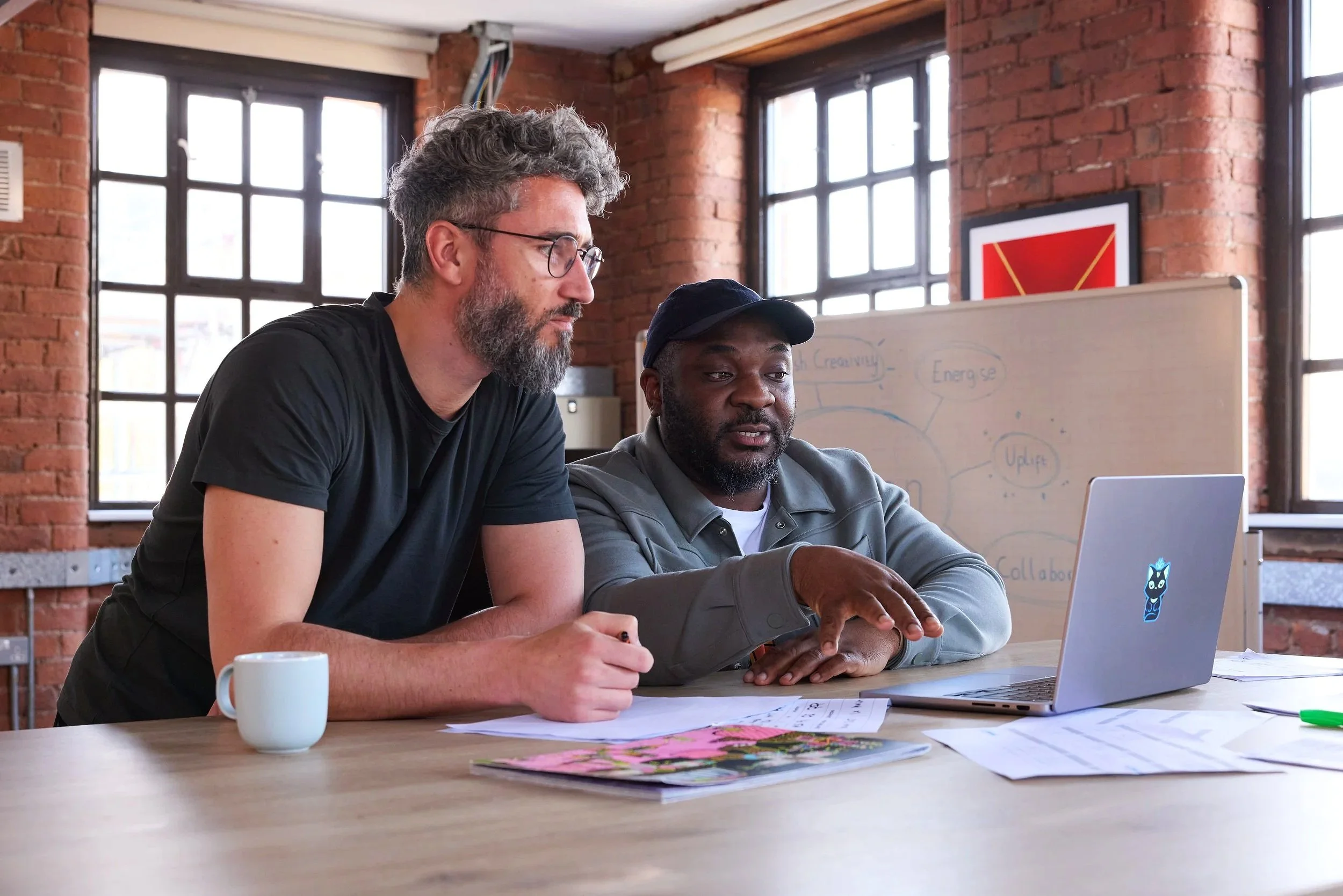 Two men working together at a desk in a bright, industrial-style office with exposed brick walls and large windows, looking at a laptop.